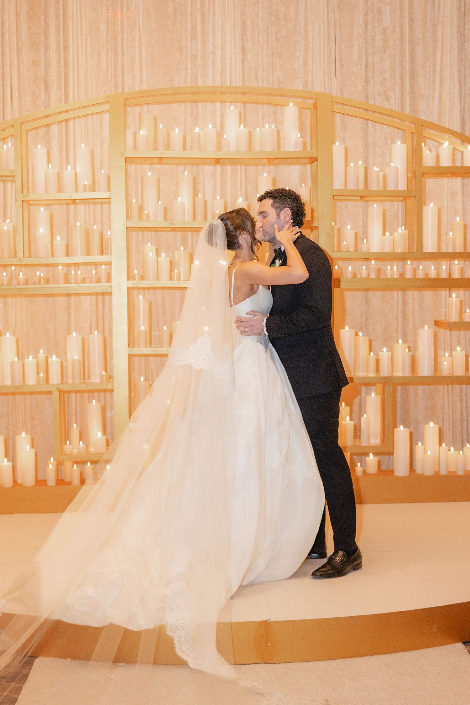 Bride and groom share first kiss at altar surrounded by elegant candlelit shelves at romantic wedding ceremony.