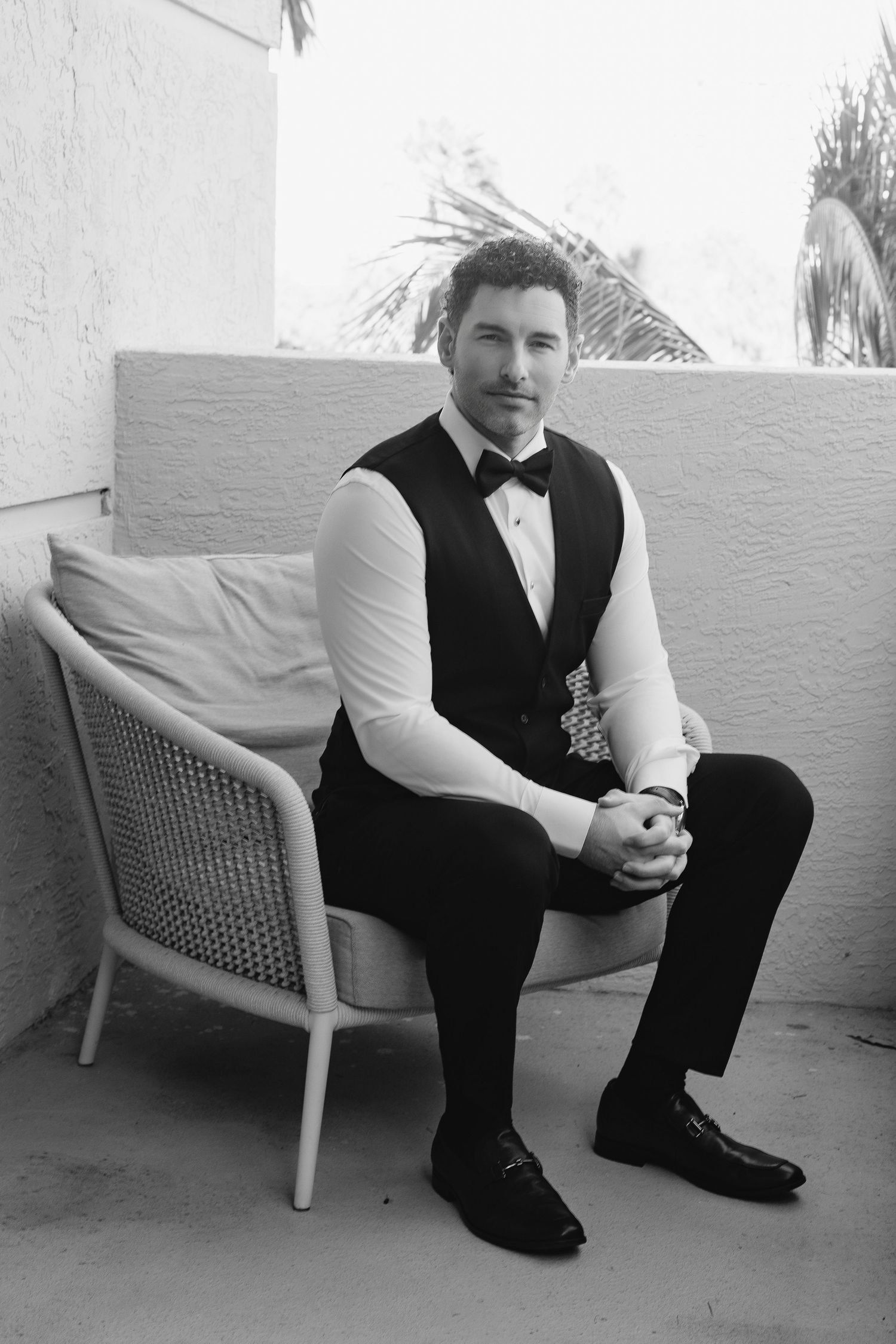 A man in a tuxedo vest and bow tie sits on a wicker chair outdoors in a black and white photo.