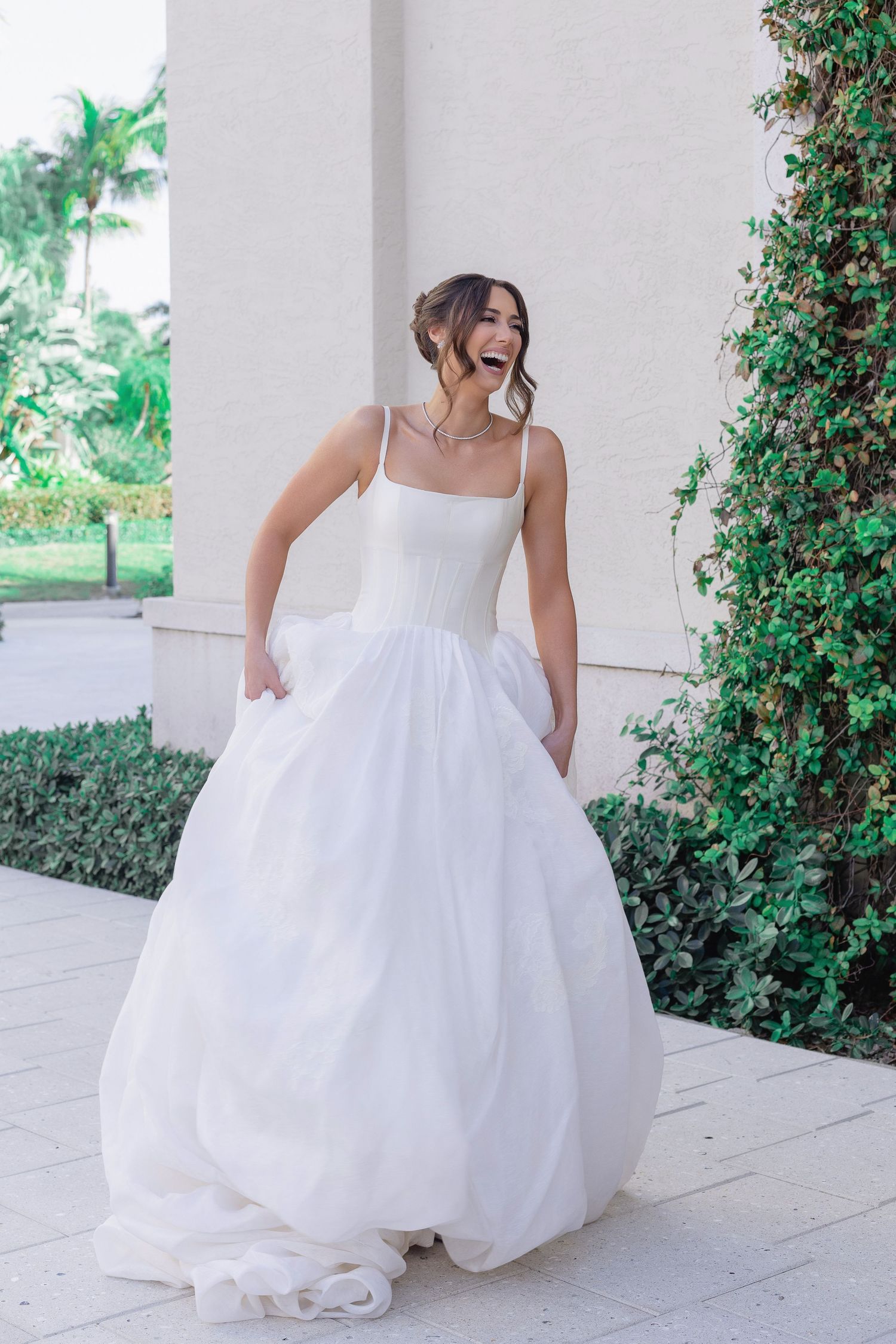 Joyful bride in a white ball gown with spaghetti straps laughs outdoors near lush greenery and a white column.
