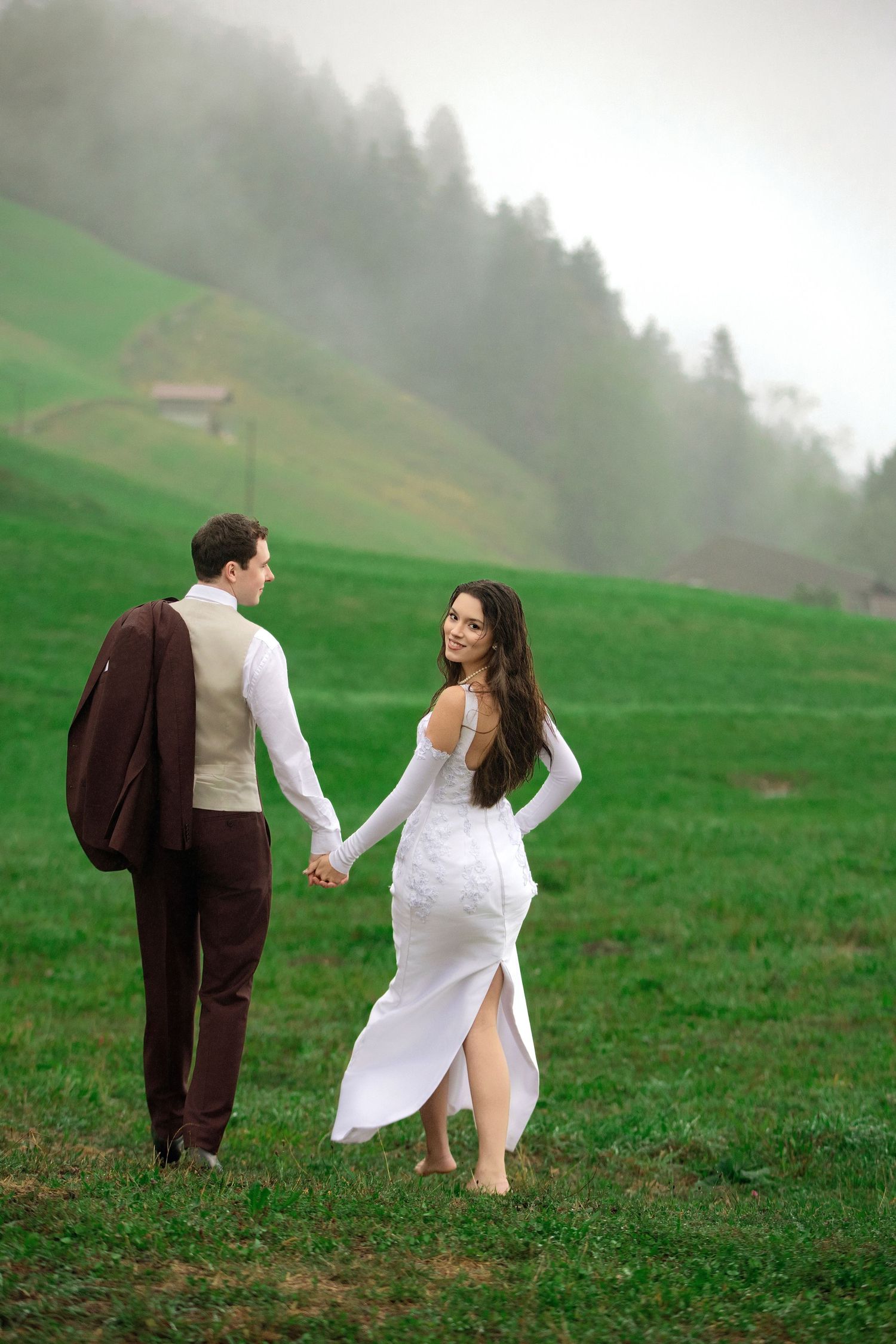 Bride and groom walking hand in hand on green misty hillside, woman in white gown glancing back smiling.