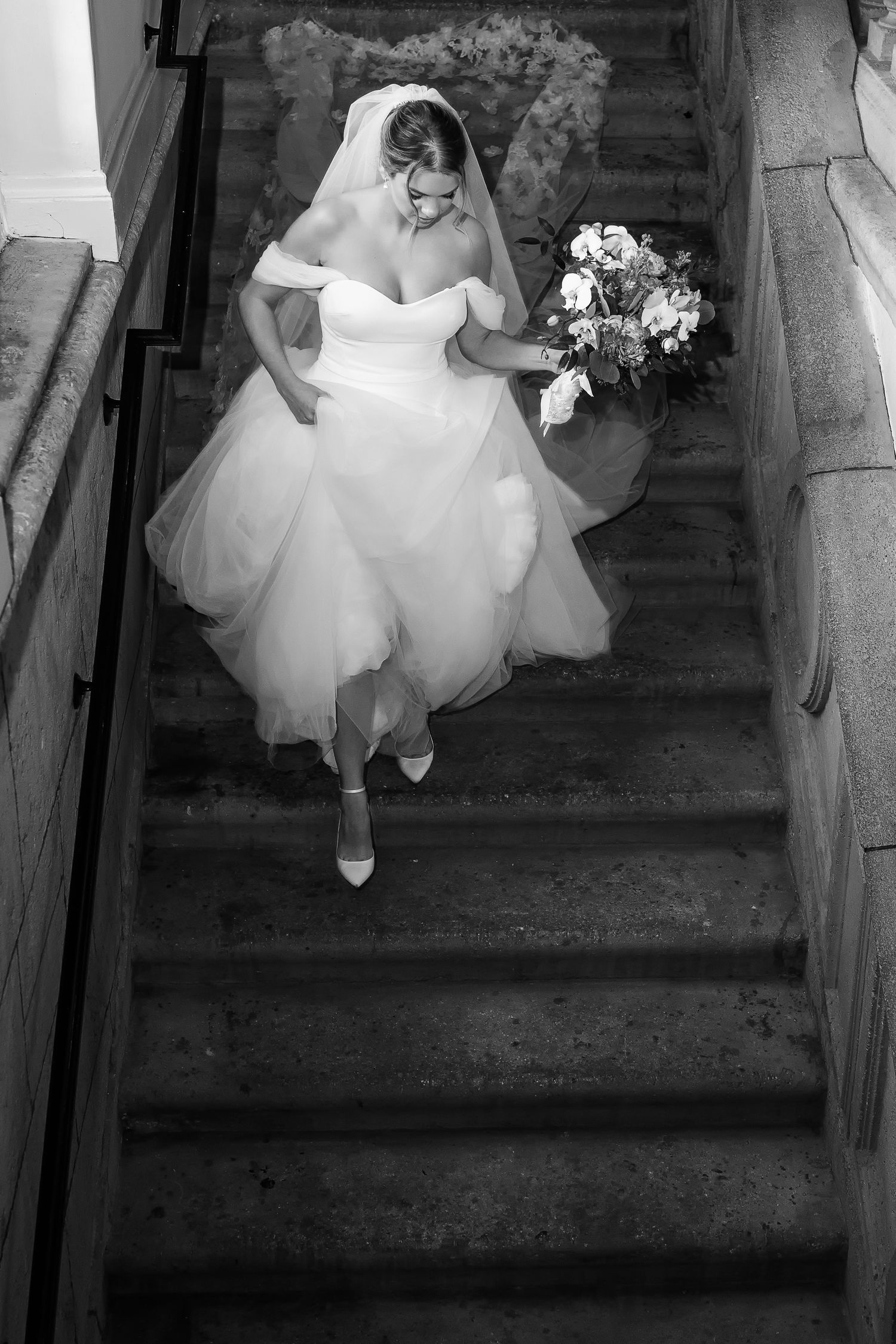 Bride in white off-shoulder gown and veil descends stone staircase holding bouquet, captured in elegant black and white.