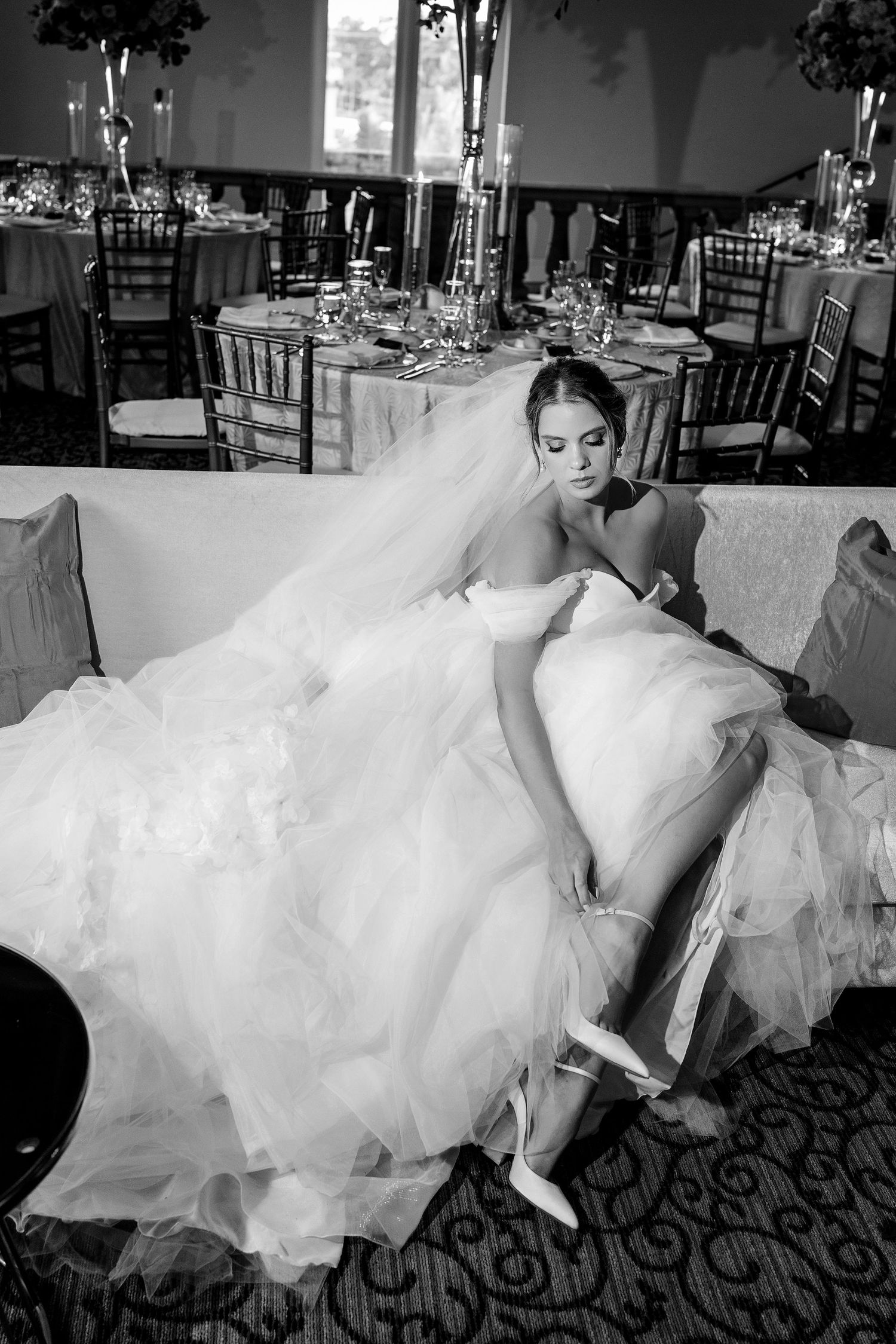 Black and white photo of a bride in a voluminous gown adjusting her heels while seated in an elegant reception hall.