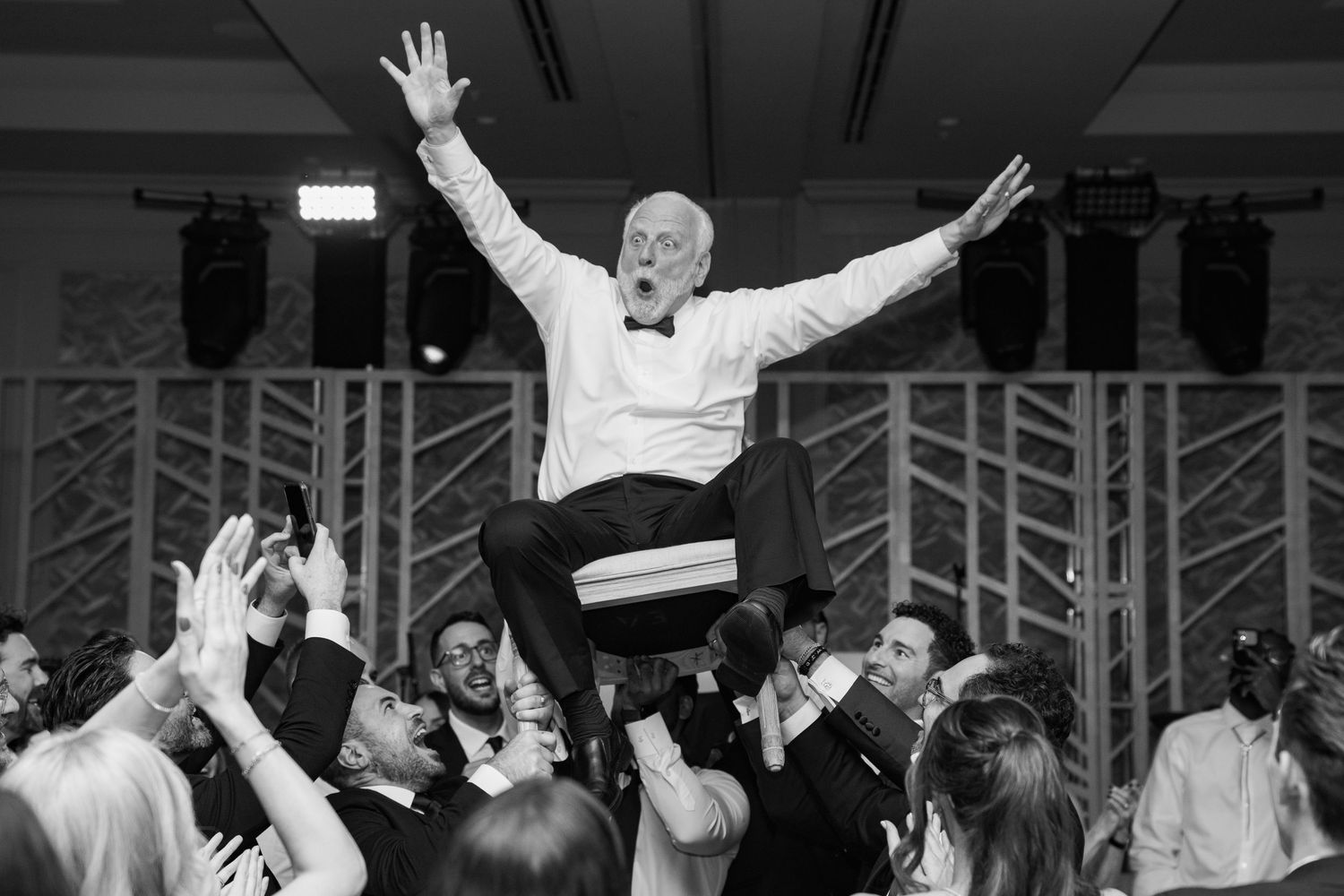 Elderly man in tuxedo joyfully lifted by crowd at wedding reception dance floor celebration.