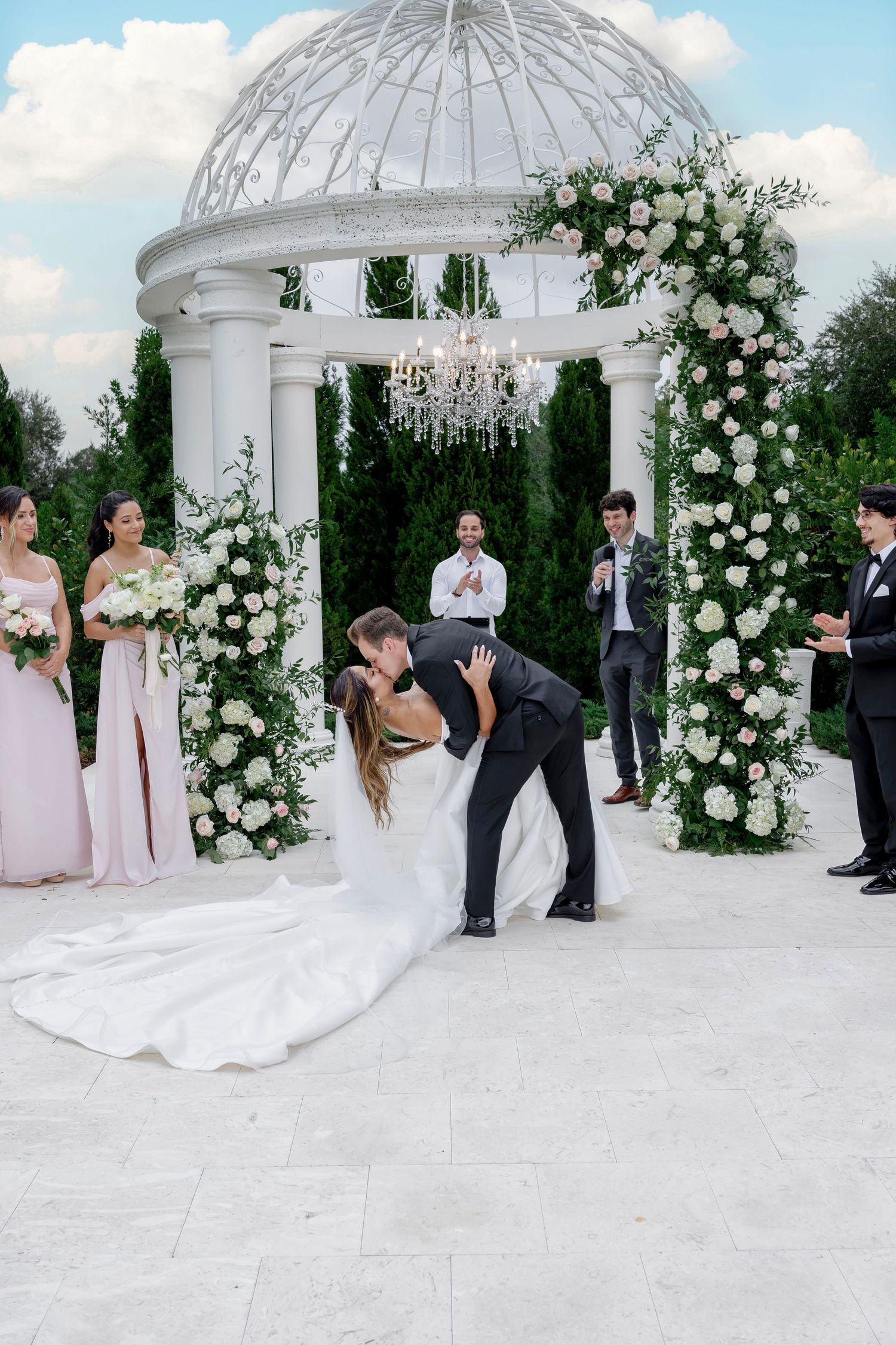 Groom dips bride for a kiss under a floral arch with white roses at an elegant outdoor wedding ceremony.
