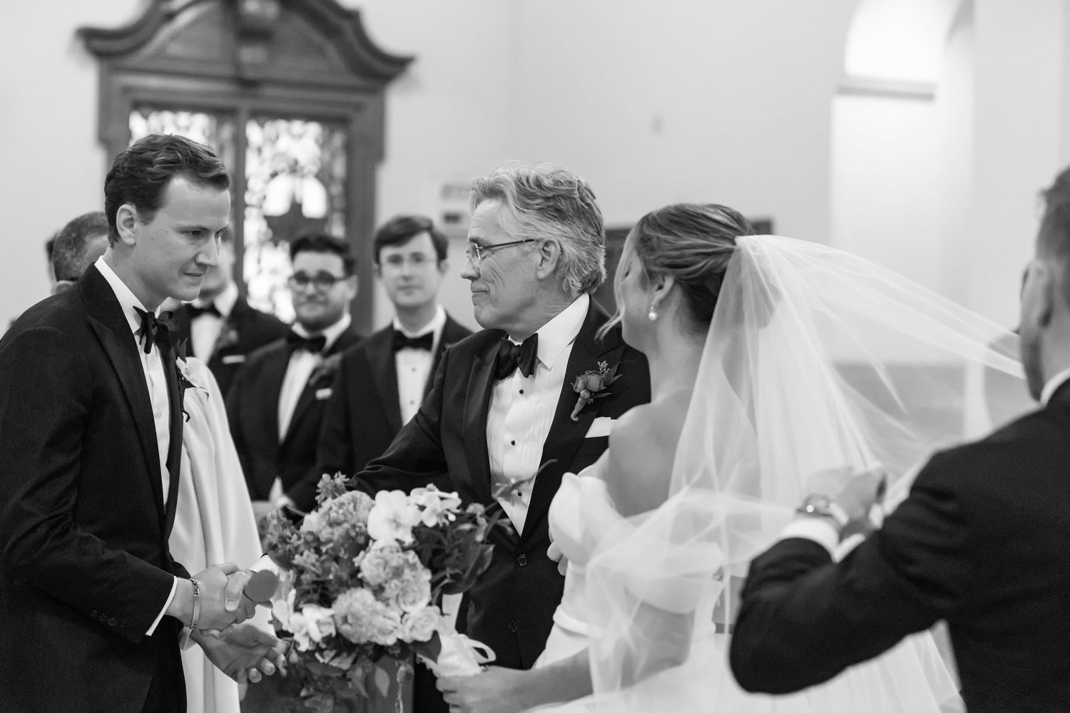 Black and white wedding ceremony photo of bride and groom greeting older gentleman in tuxedo at elegant church venue.
