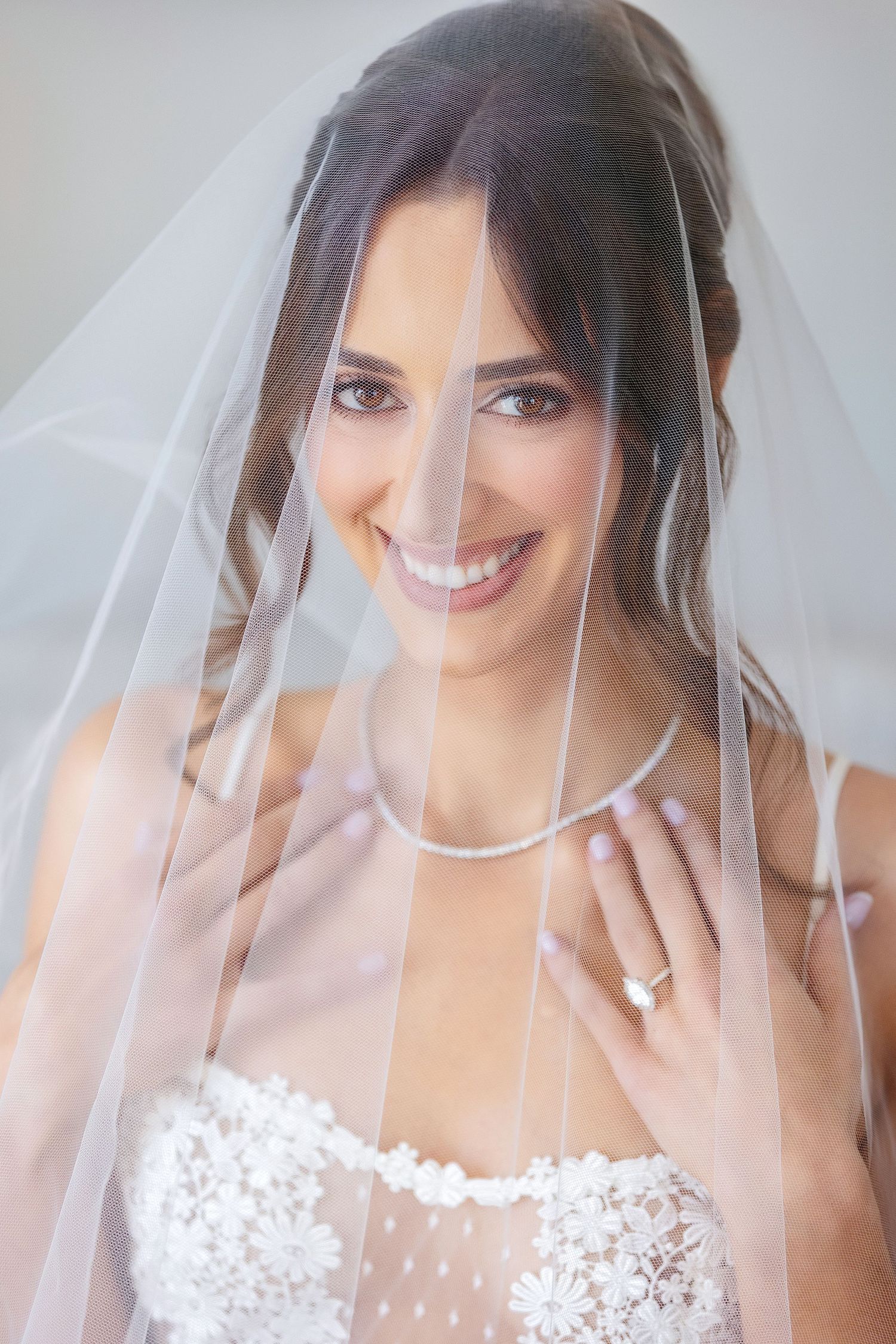 Smiling bride in lace wedding dress with veil draped over her, wearing a diamond necklace and engagement ring.