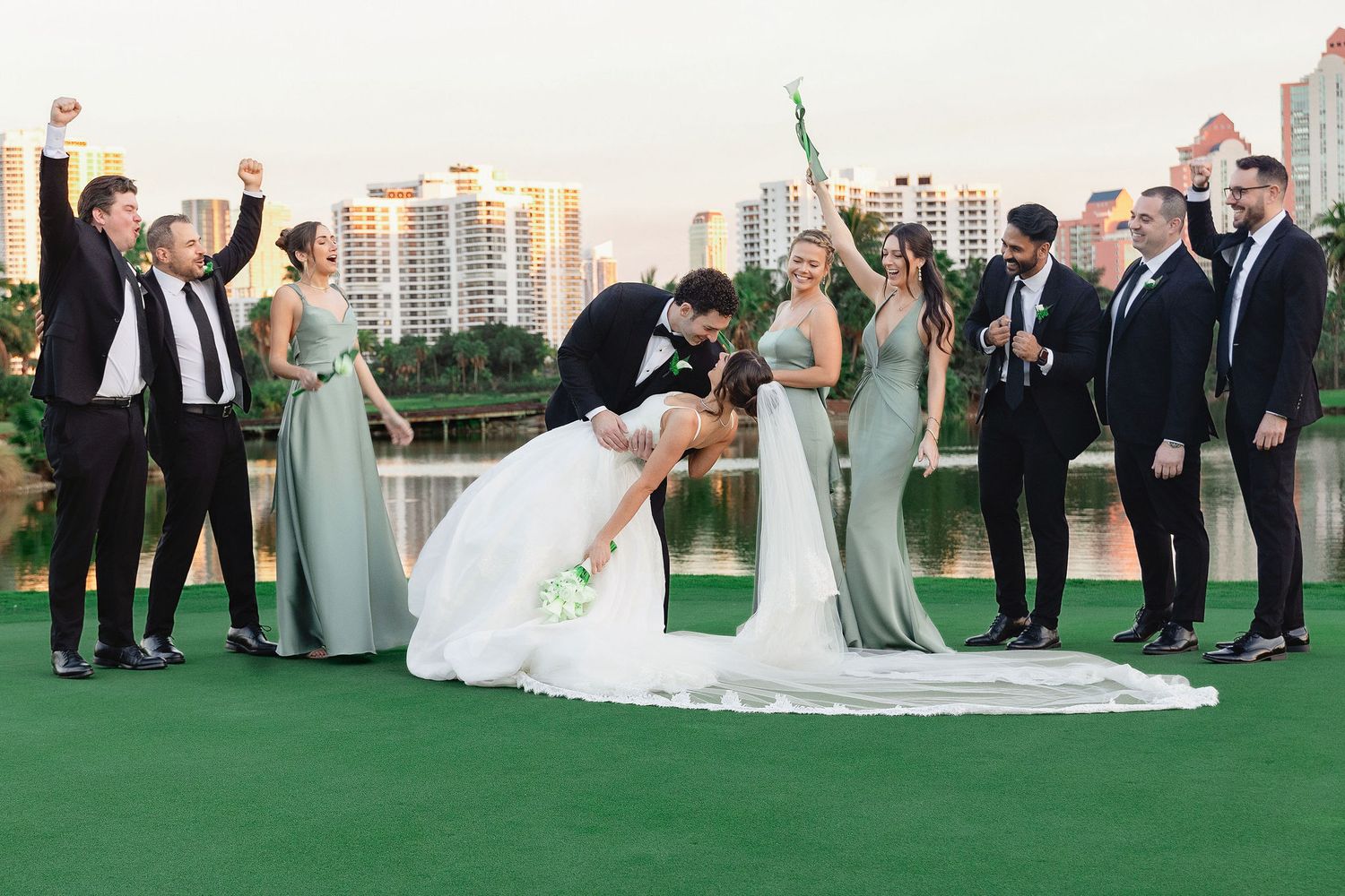 Wedding party celebrates on a green rooftop venue with city skyline in the background at sunset.