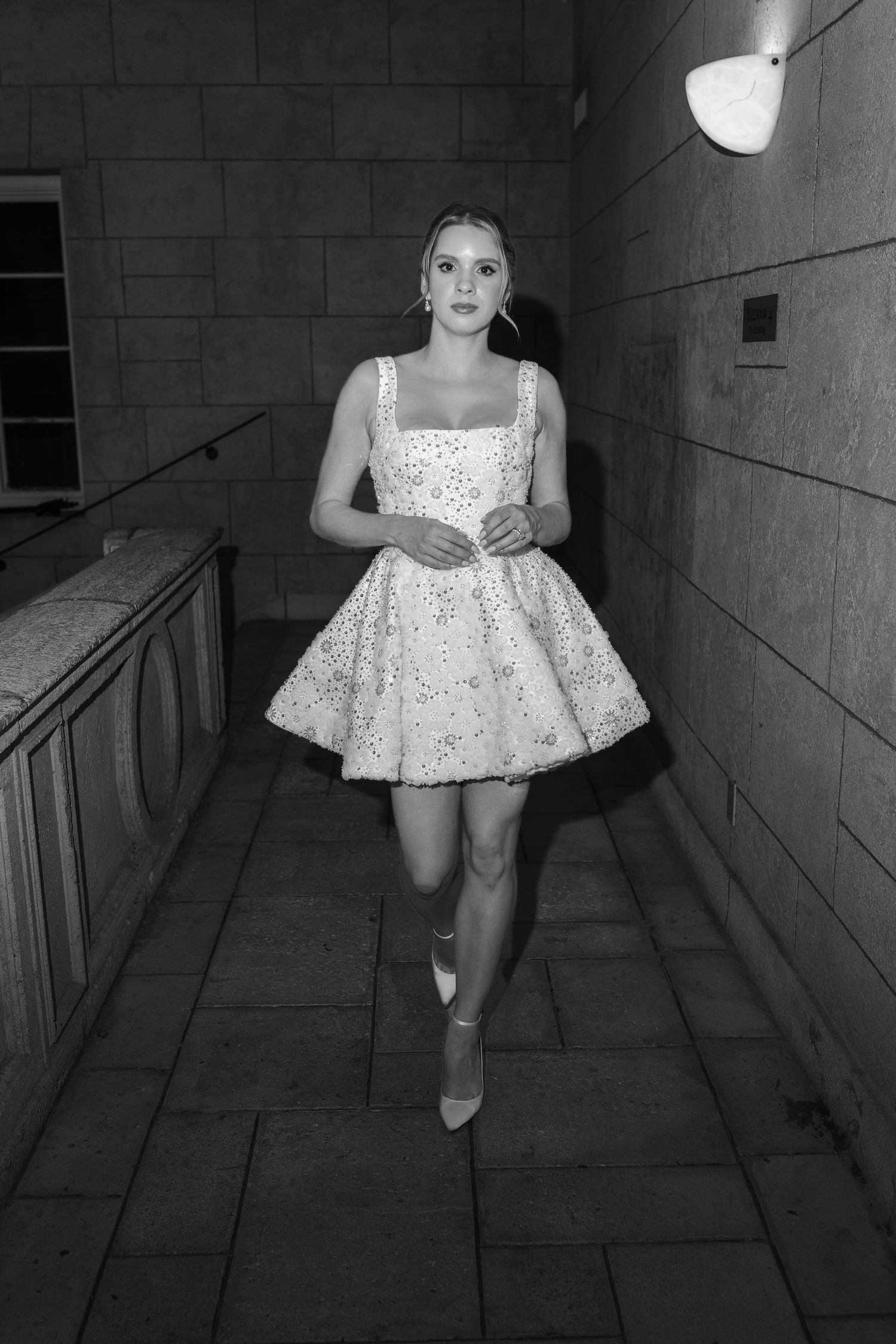 Black and white photo of a young woman in a floral mini dress and heels walking through a stone corridor at night.