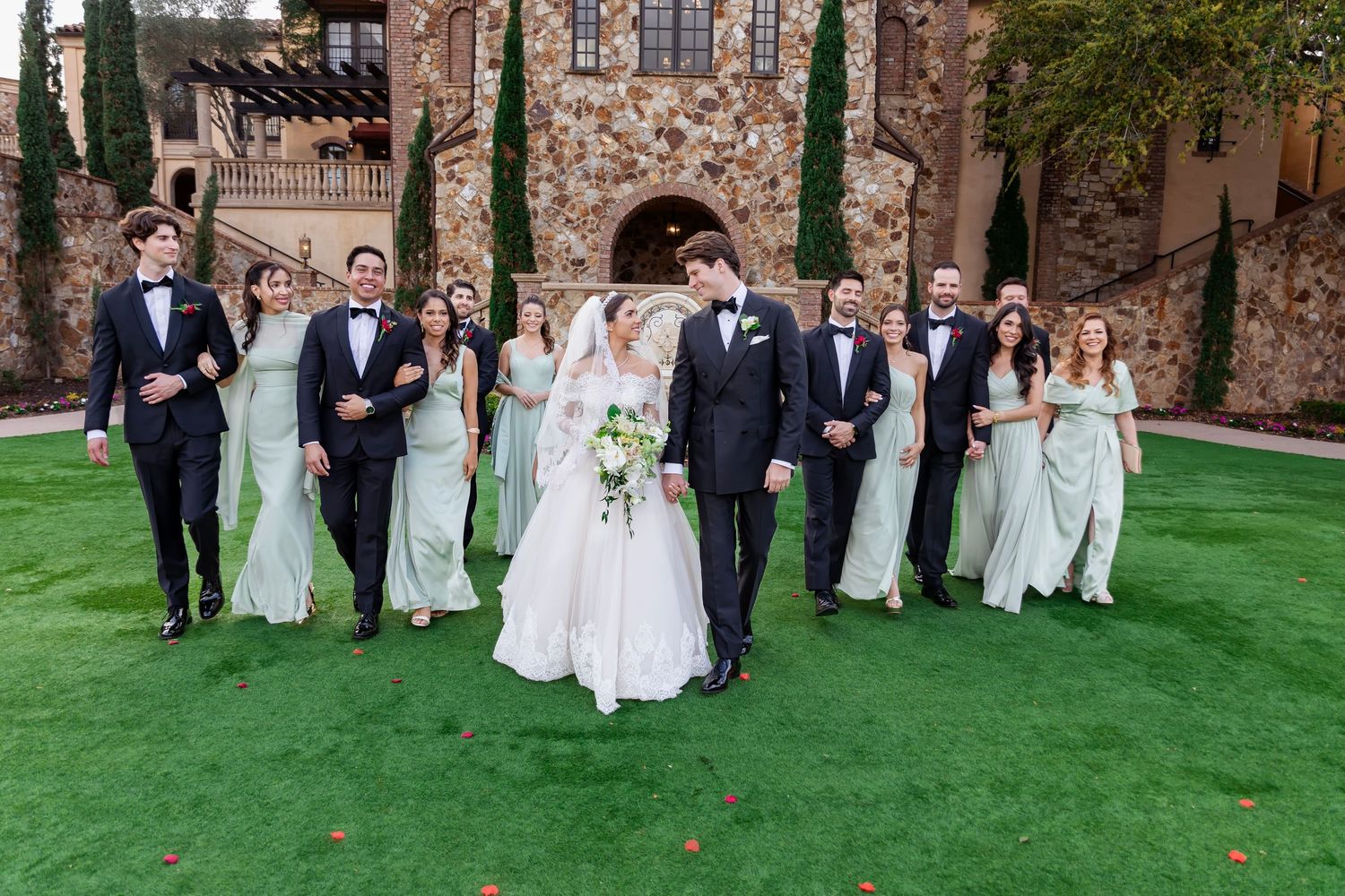 Wedding party in formal attire poses on manicured lawn in front of stone building.