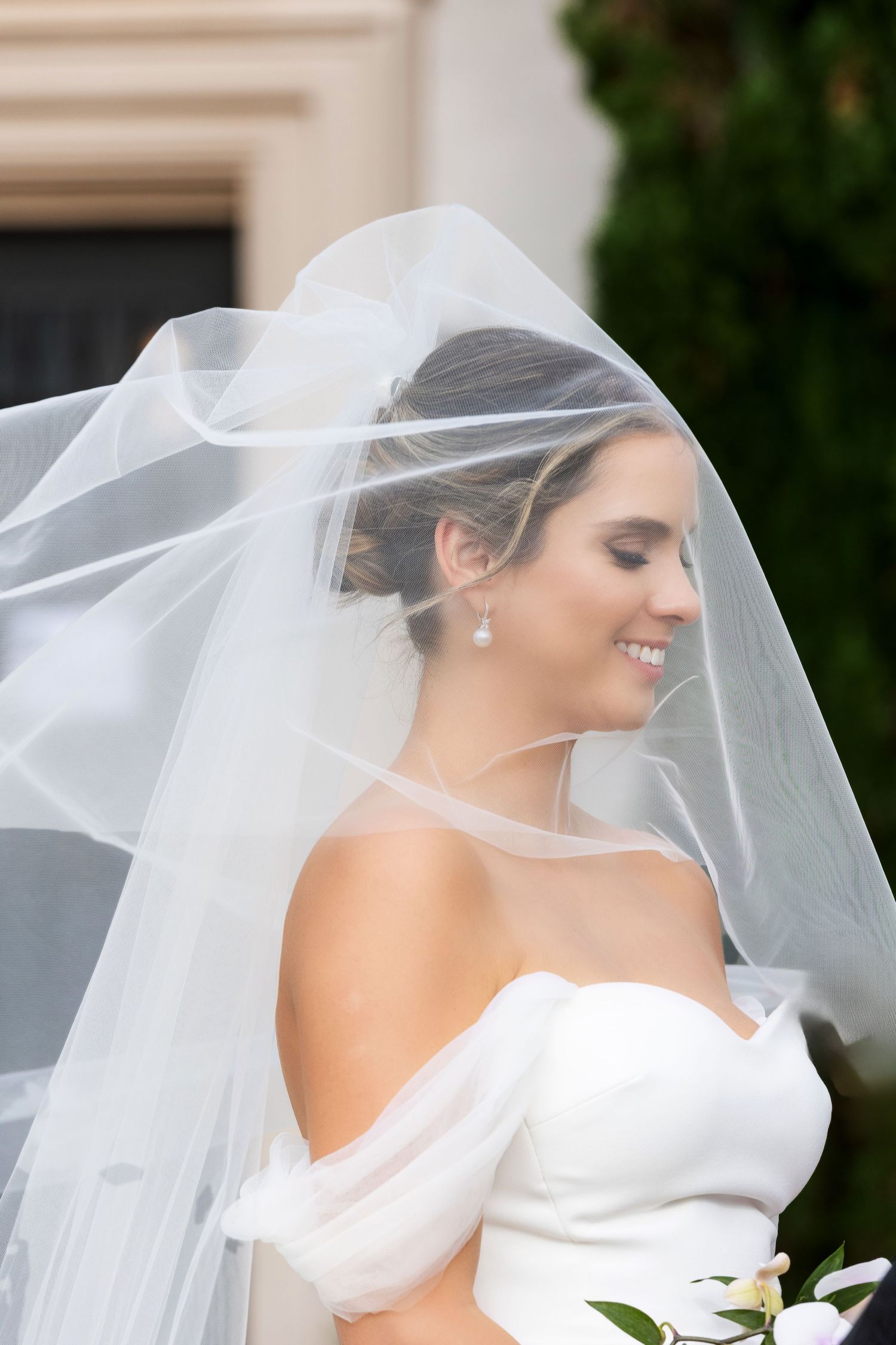 Smiling bride in a strapless white gown with flowing veil and pearl earrings on her wedding day.