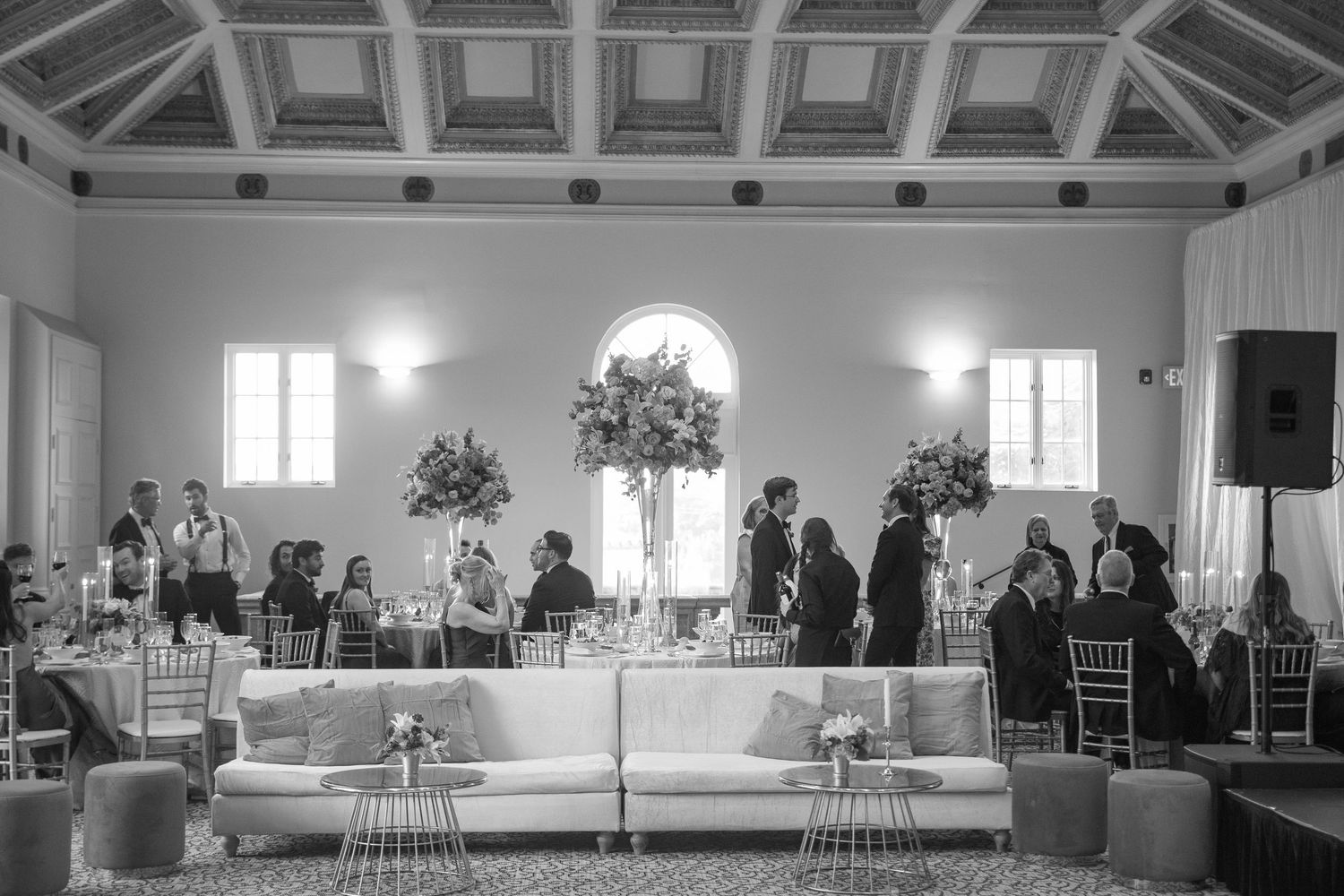 Black and white photo of an elegant wedding reception hall with floral arrangements, guests mingling, and a white sofa seating area.