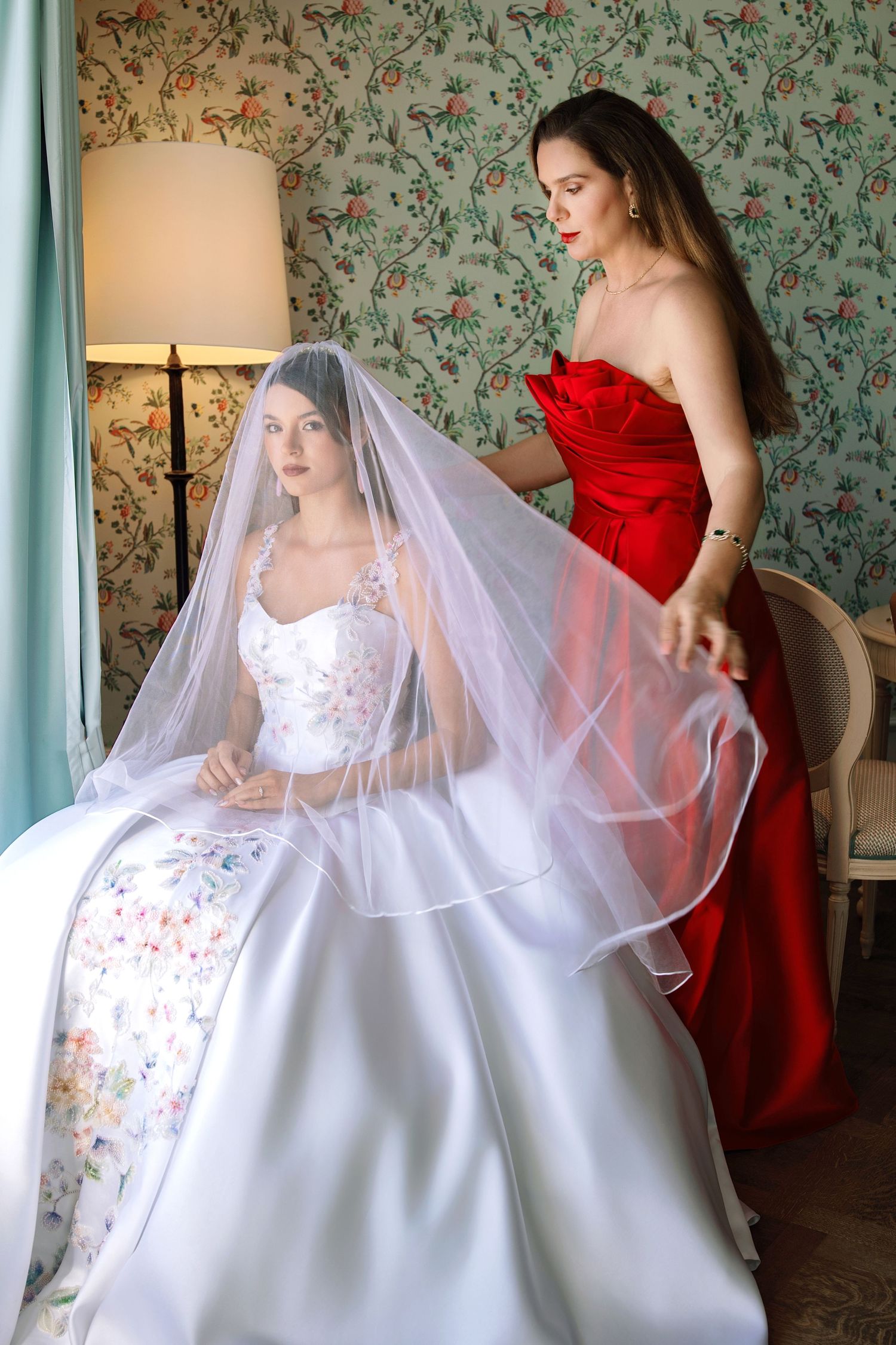 A bride in a floral wedding gown and veil sits as a bridesmaid in a red strapless dress adjusts her veil.