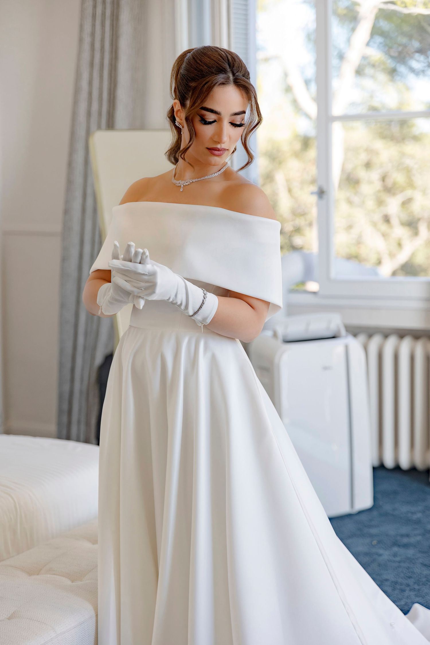 Elegant bride in off-shoulder white wedding gown and long gloves stands gracefully in sunlit room with classic decor.