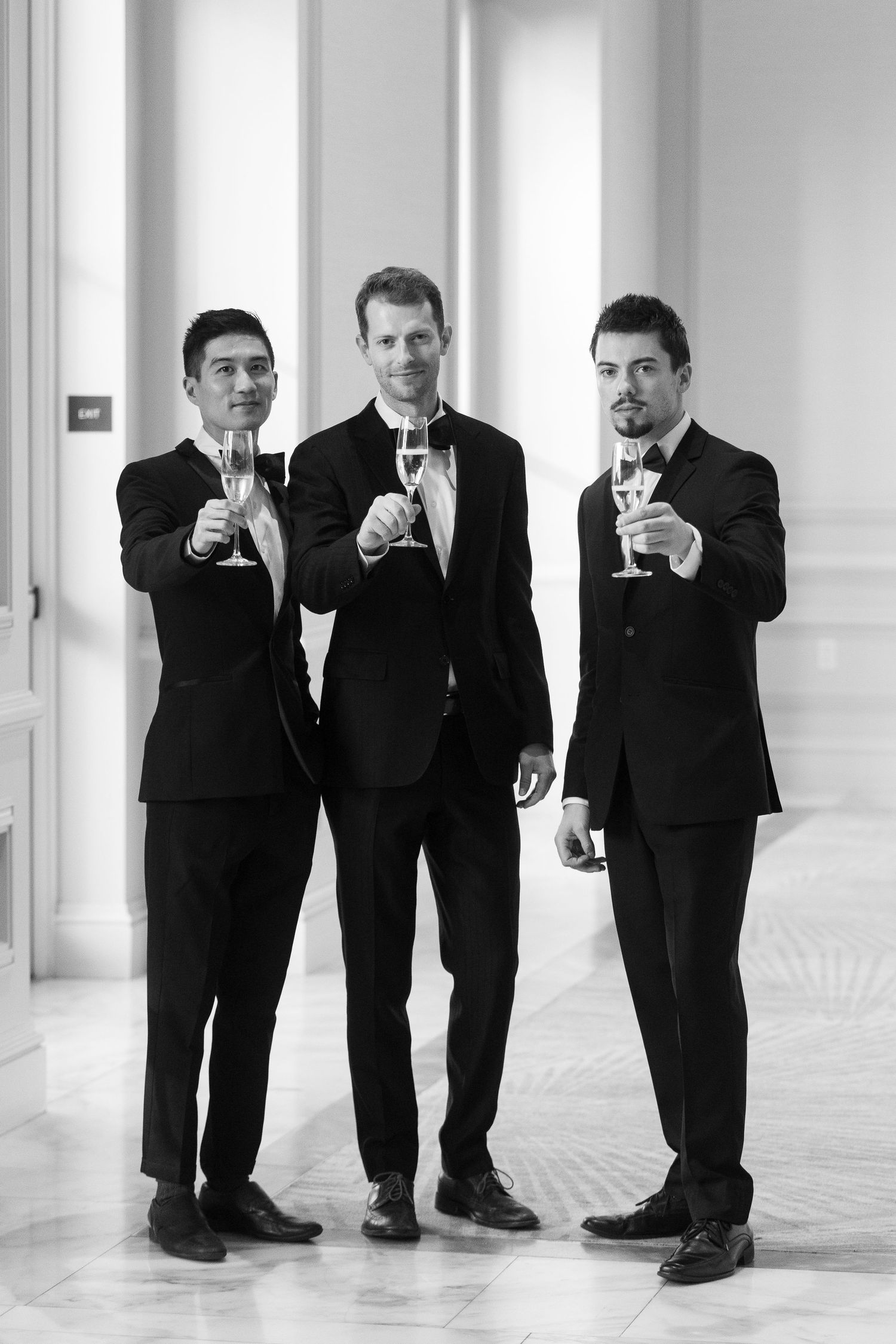 Three men in black suits toasting with champagne glasses in a elegant hall, black and white photo.