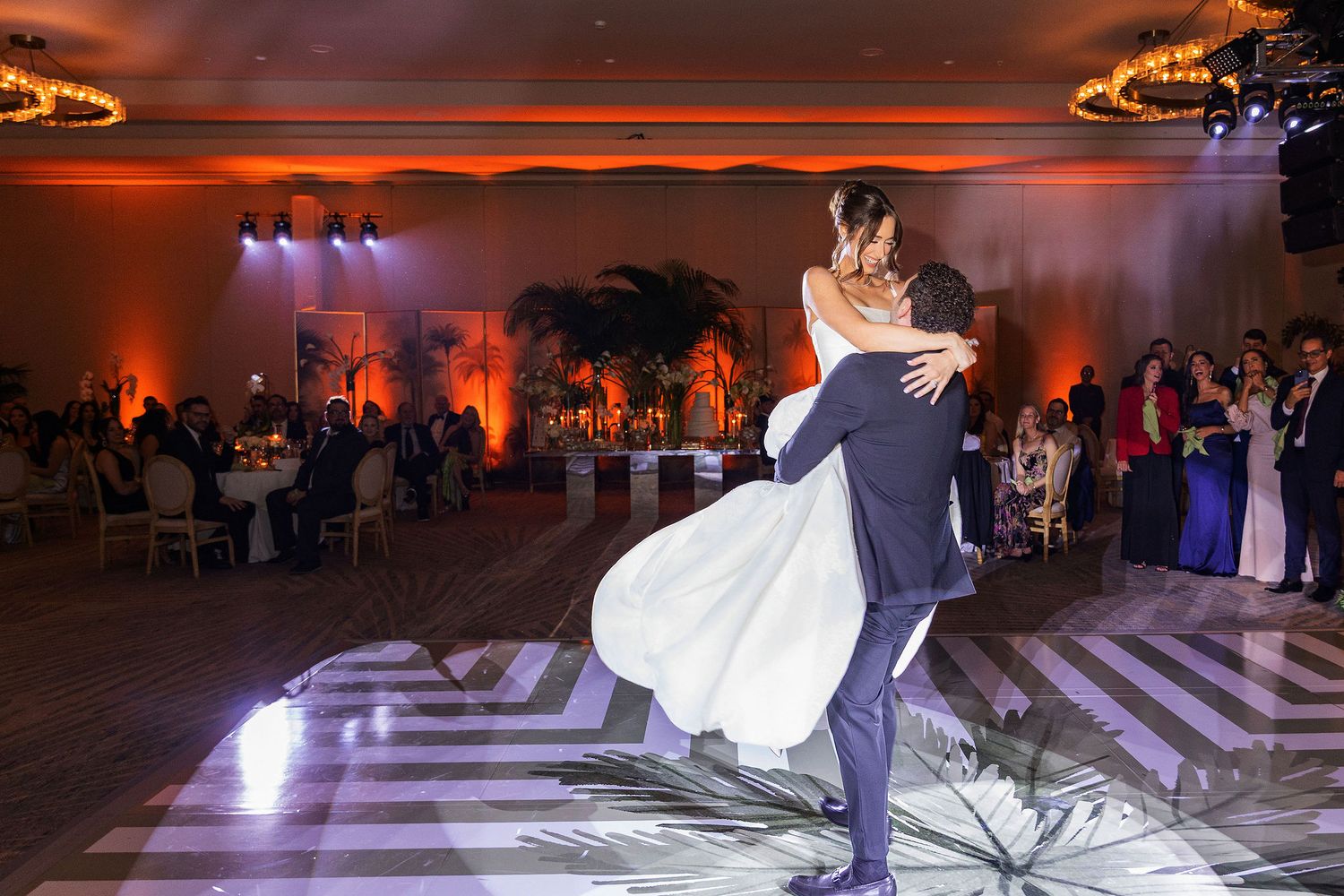 A couple shares their first dance at a wedding reception with dramatic orange lighting and a patterned dance floor.