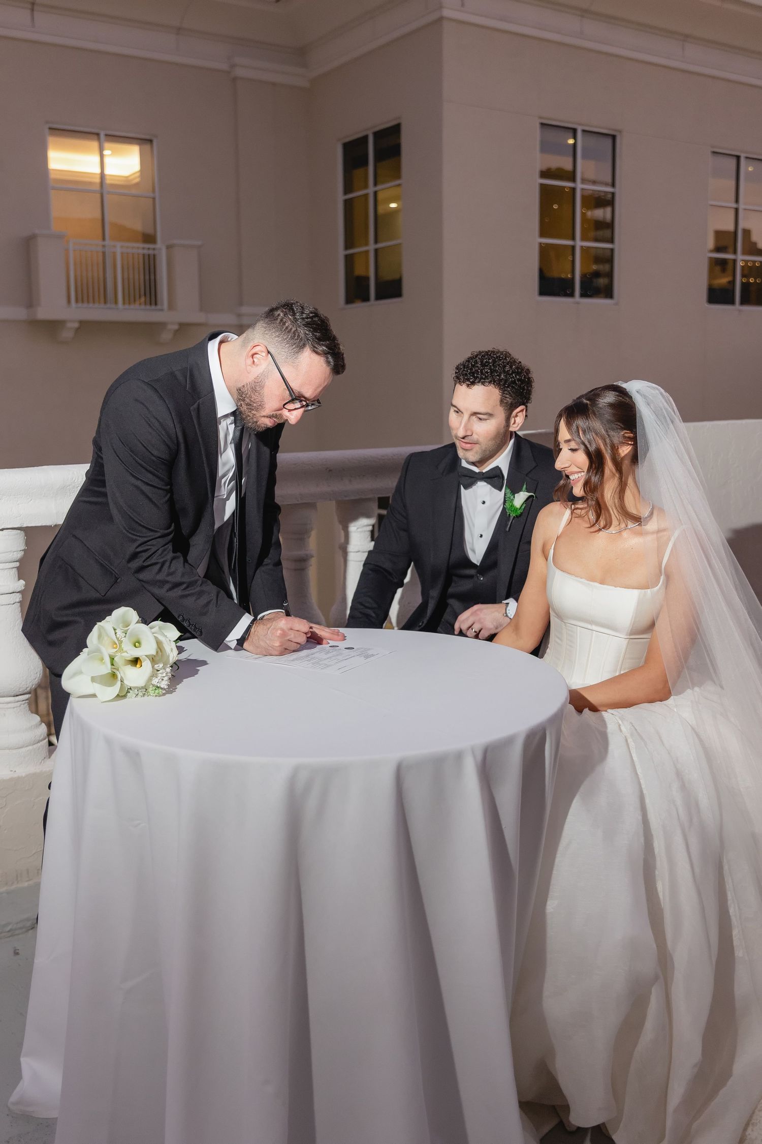 Bride and groom signing wedding documents with officiant at elegant white table during evening ceremony.