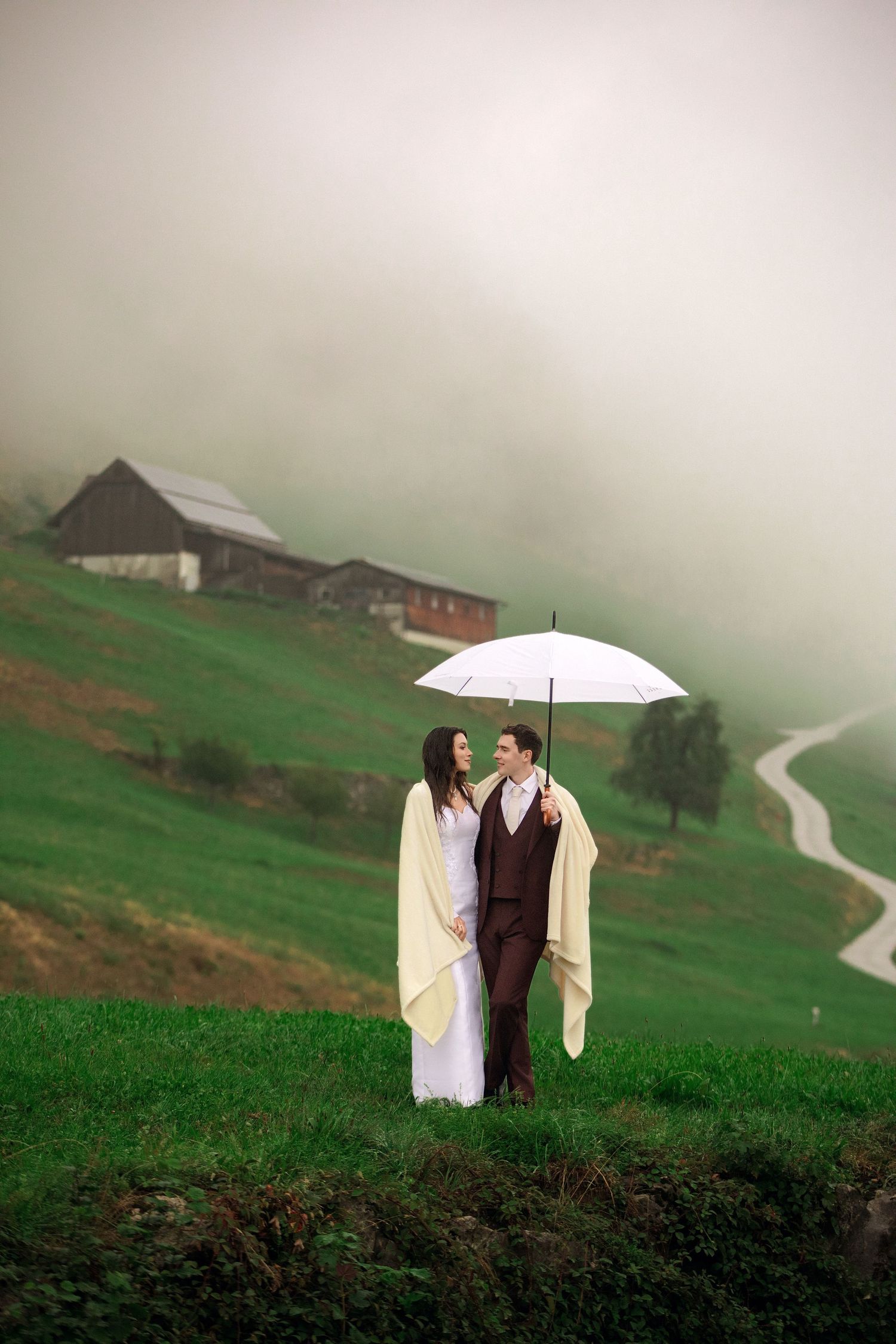 Couple sharing a white umbrella on a misty green hillside, with a rustic barn and winding path in the background.