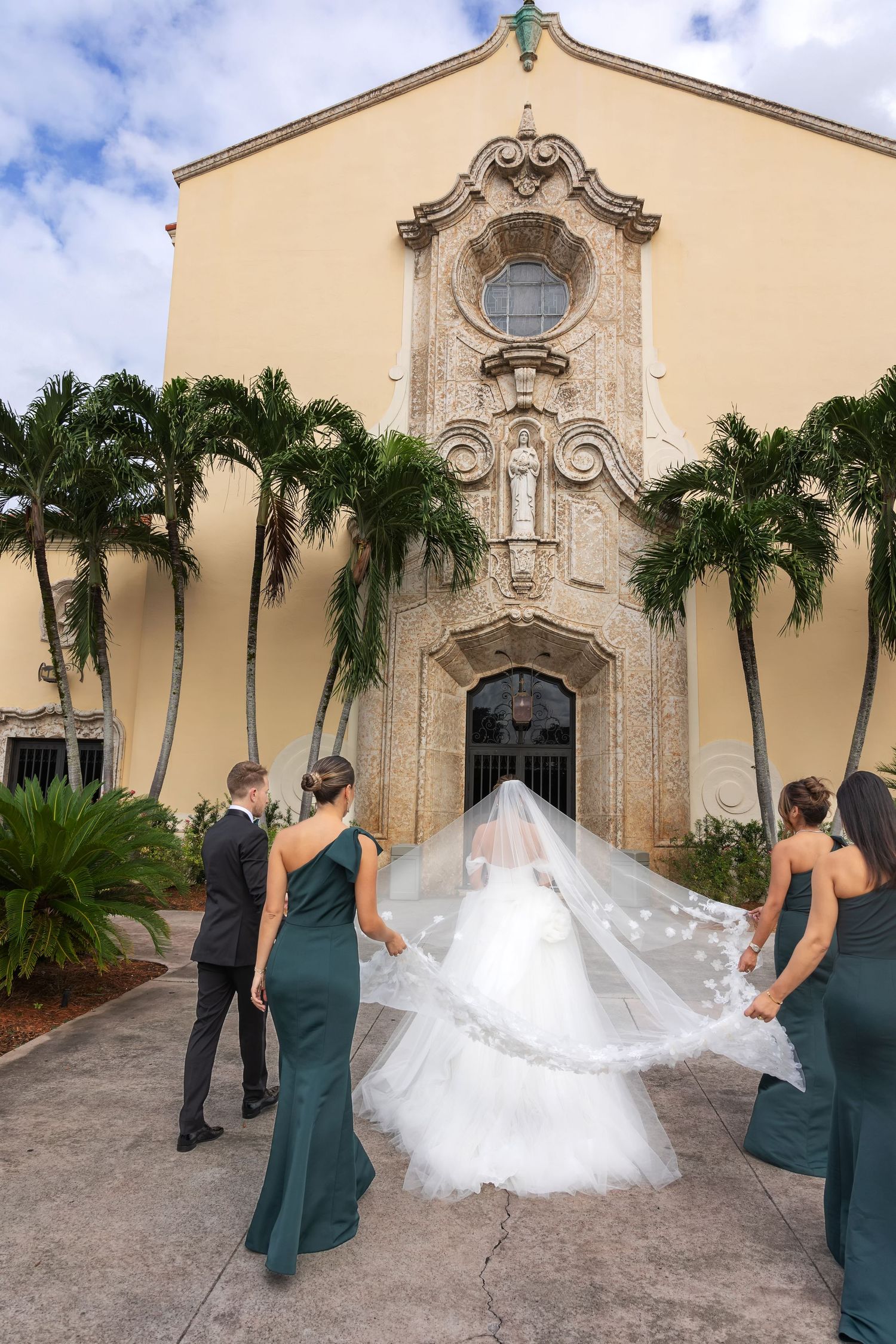 Bride in white gown with flowing veil walks toward yellow church entrance, flanked by bridesmaids in green dresses.