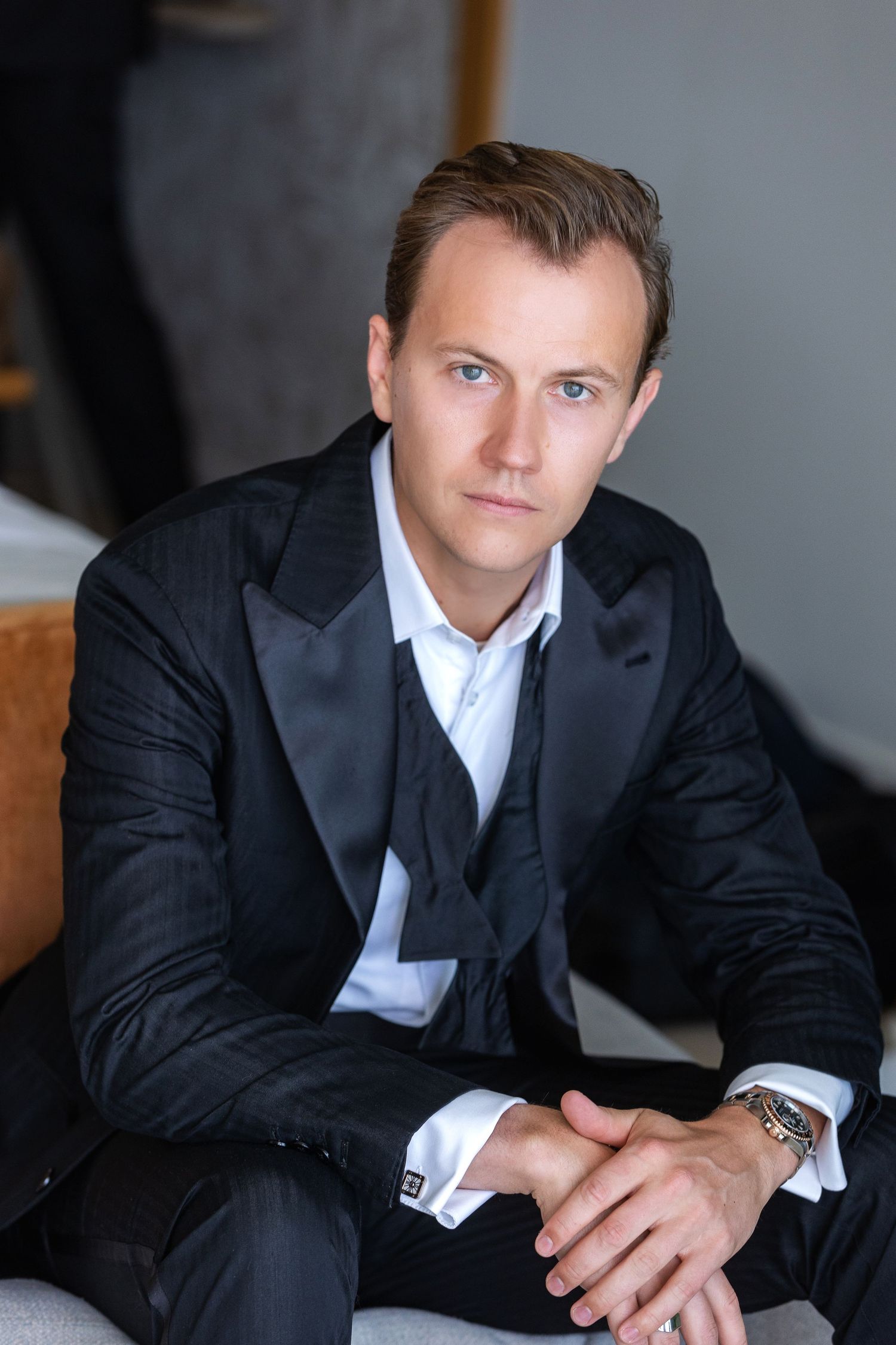 Elegant man in black tuxedo with white shirt sits confidently, looking at camera in a sophisticated portrait.