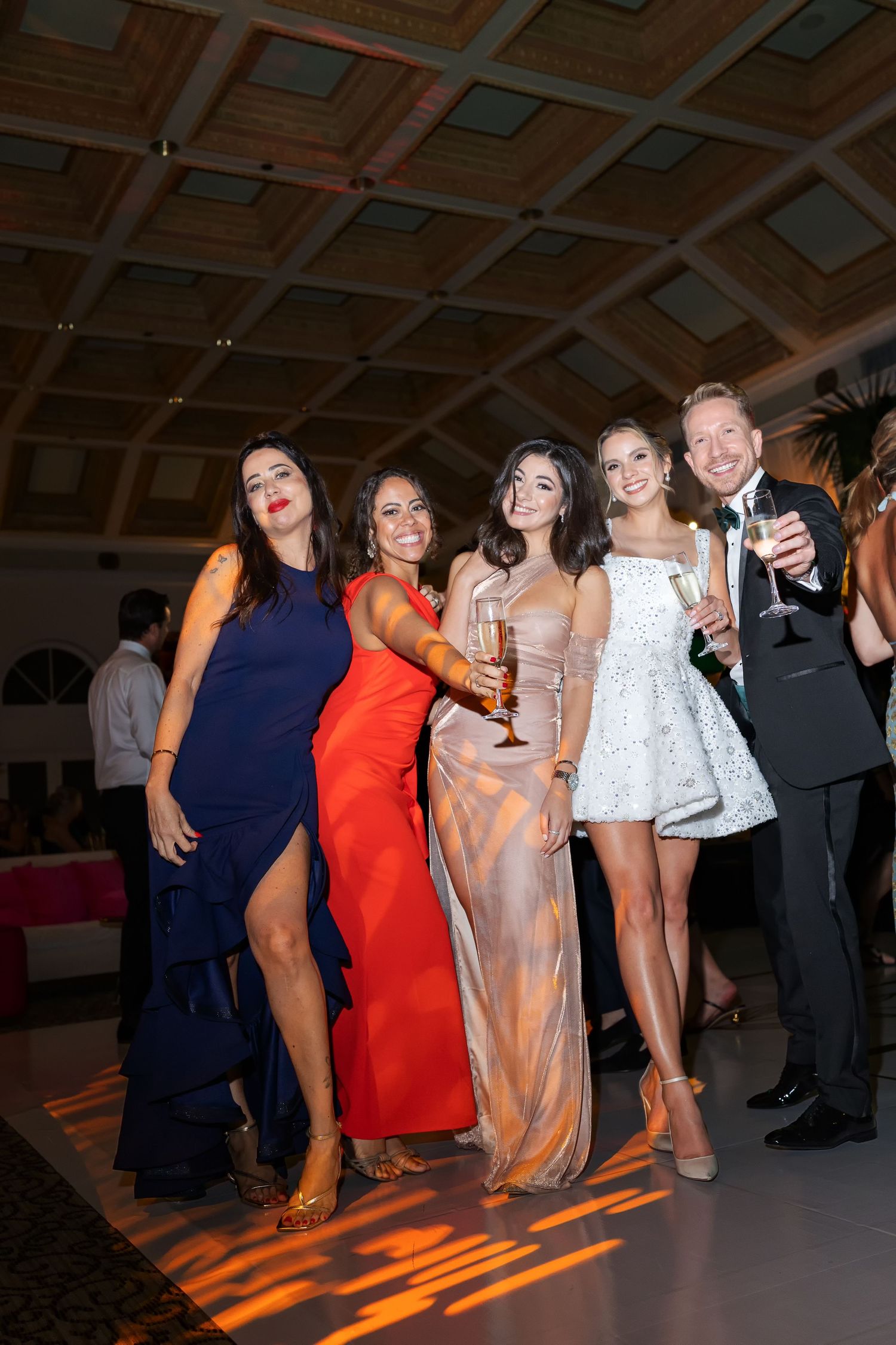 Group of elegantly dressed friends posing with champagne glasses at a formal gala event in a grand ballroom.