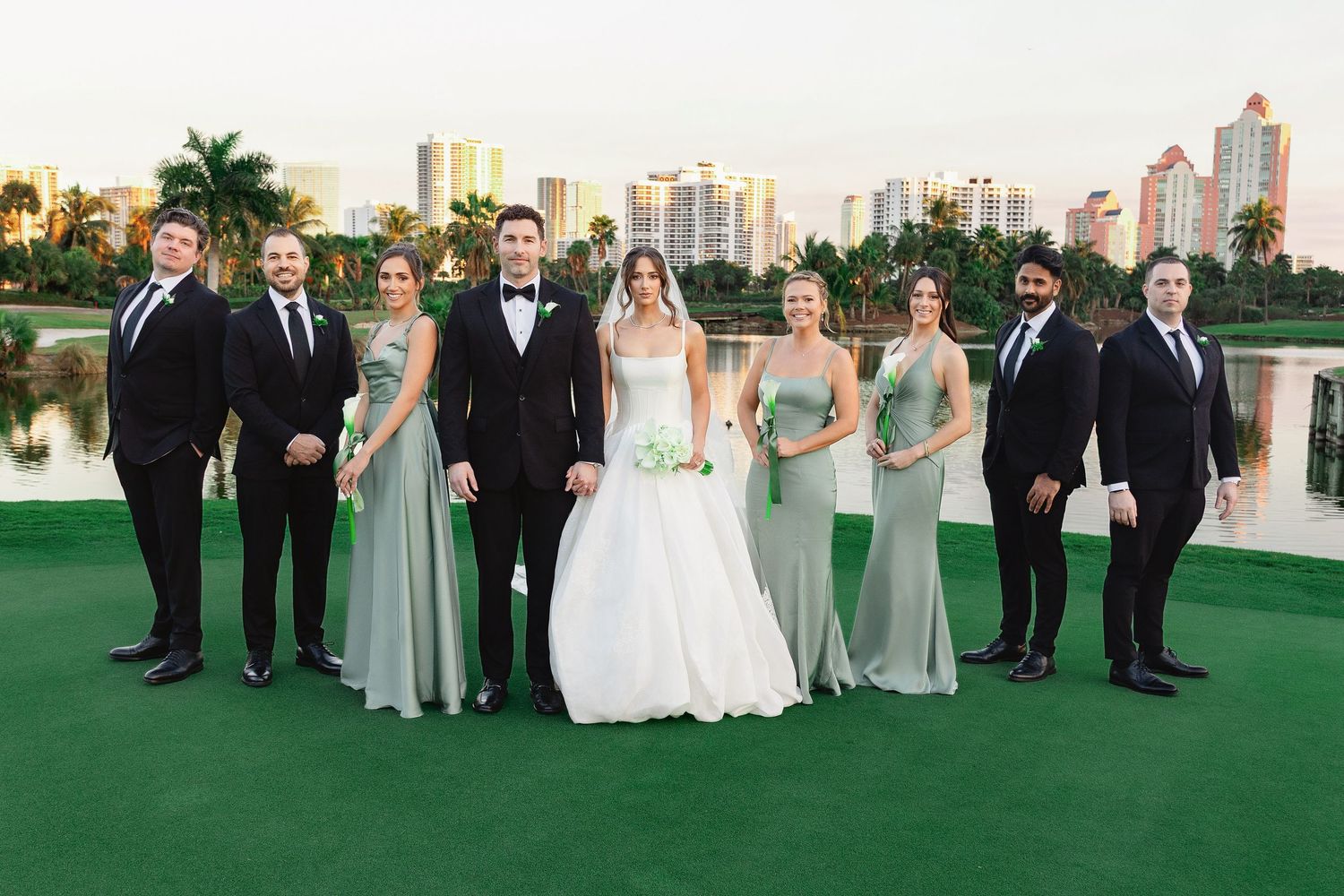 Wedding party poses outdoors on green golf course with city skyline, bridesmaids in sage gowns, groomsmen in black suits.