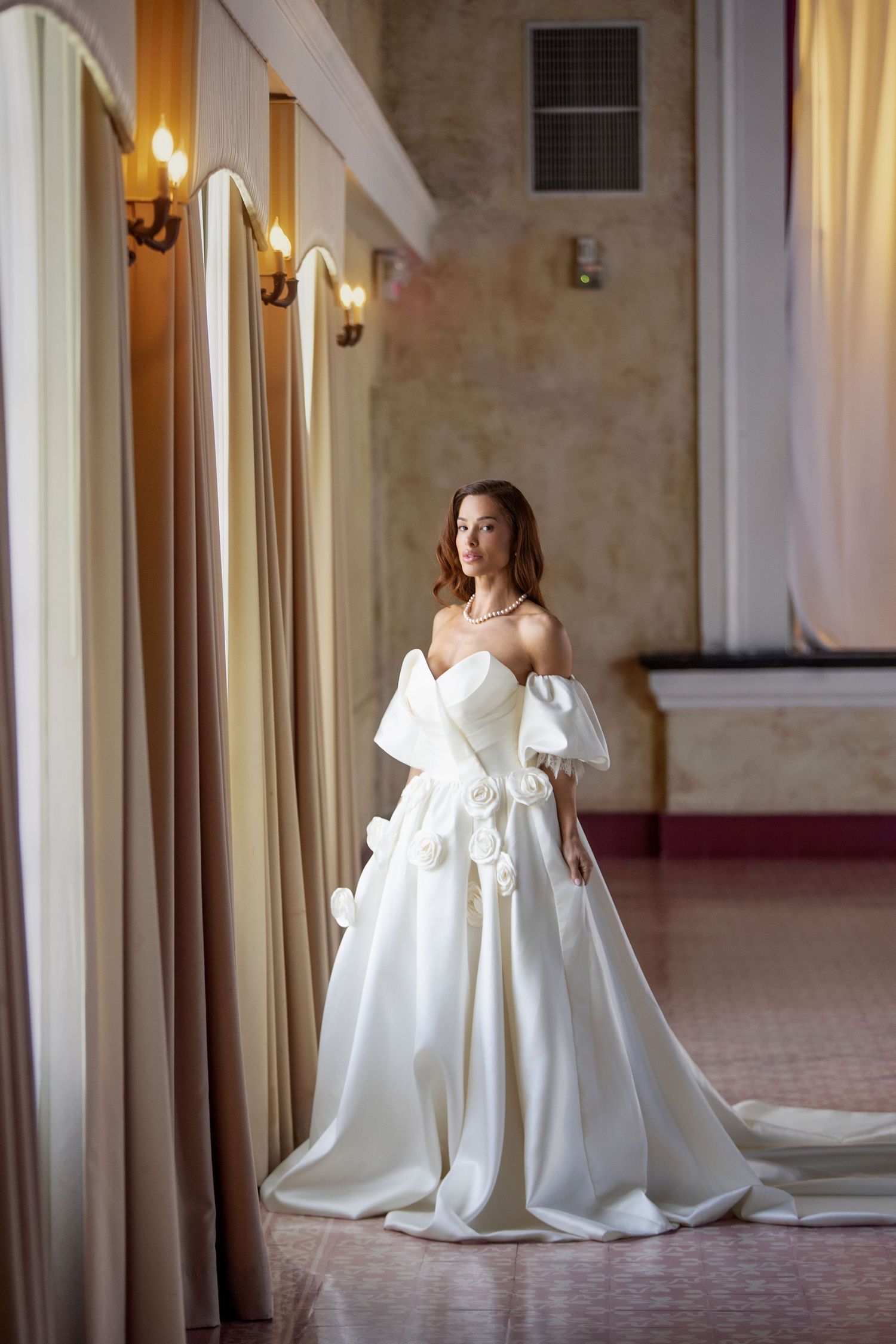 Elegant bride in a white off-shoulder ball gown with floral details poses in a grand hall.