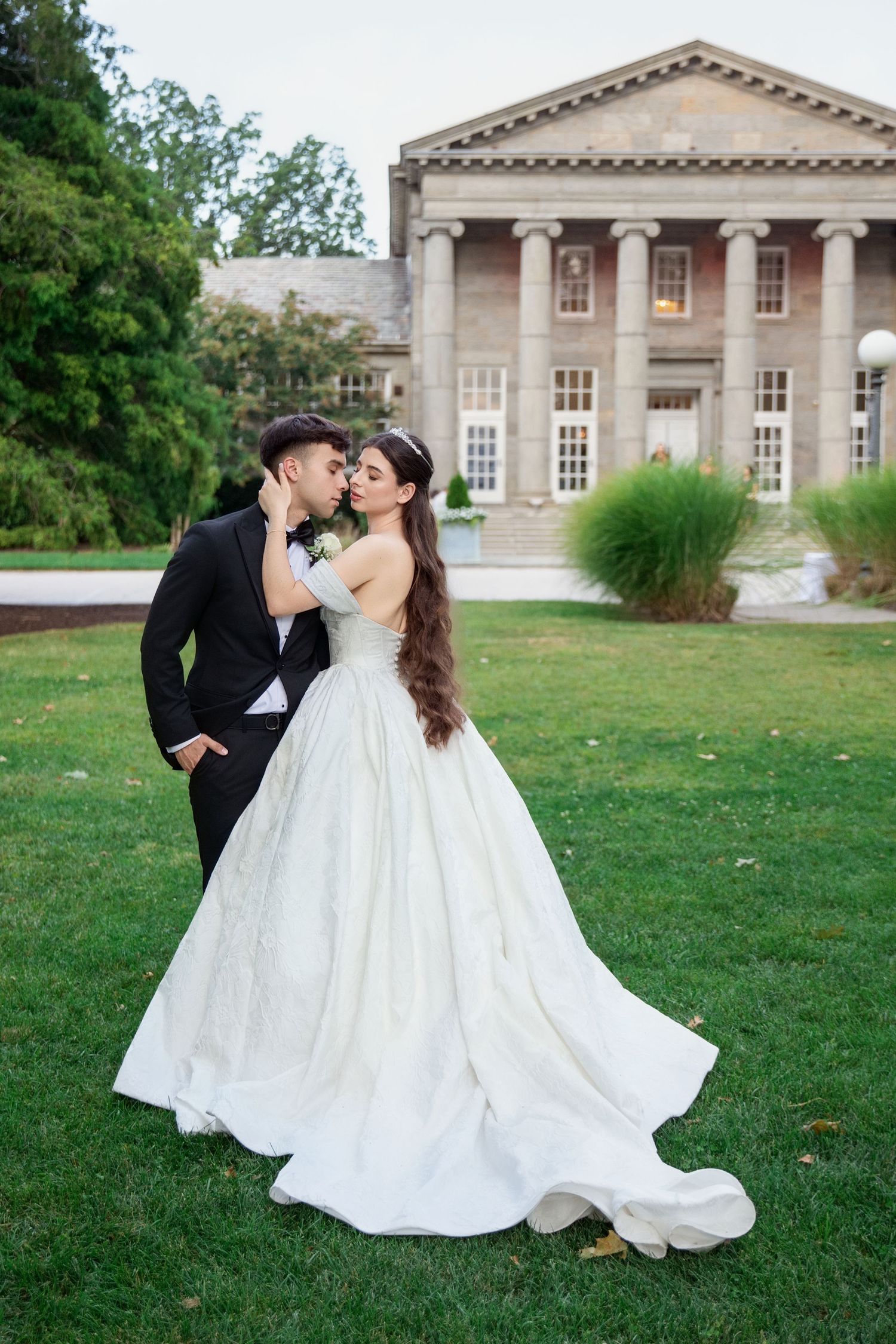 A romantic couple in wedding attire dances and embraces on a grand estate lawn with neoclassical architecture in background.