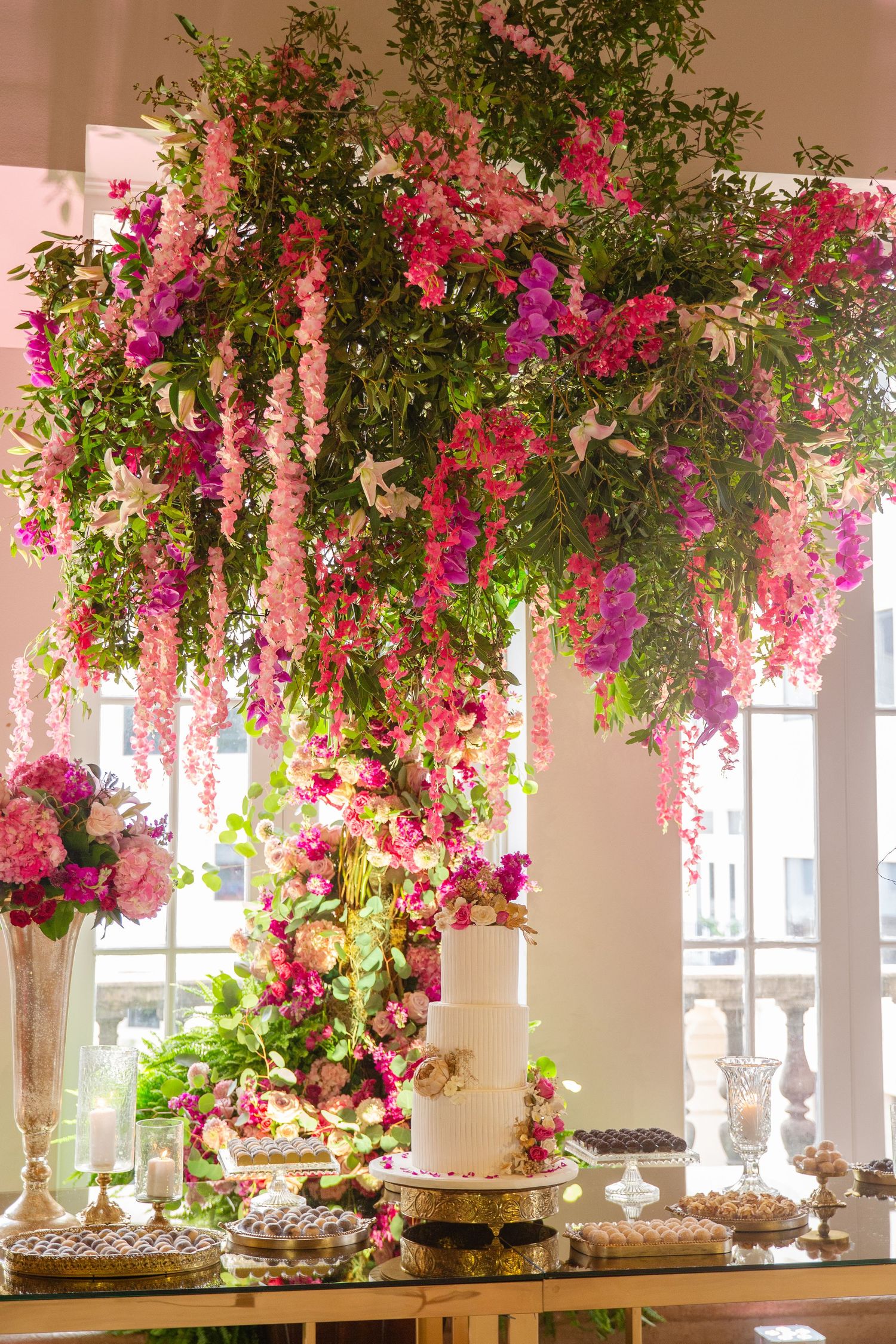 Lush pink and magenta floral installation cascades over an elegant wedding dessert table with a white tiered cake.