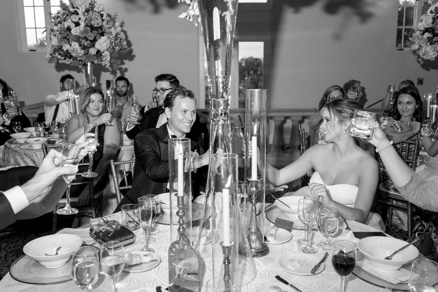 Black and white photo of bride and groom toasting with guests at elegant wedding reception dinner table.