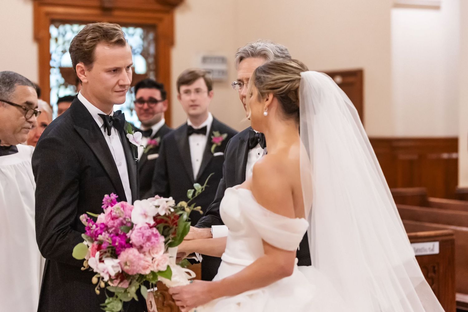 Bride and groom exchange vows at church altar, bride holding pink bouquet in elegant white off-shoulder gown and veil.