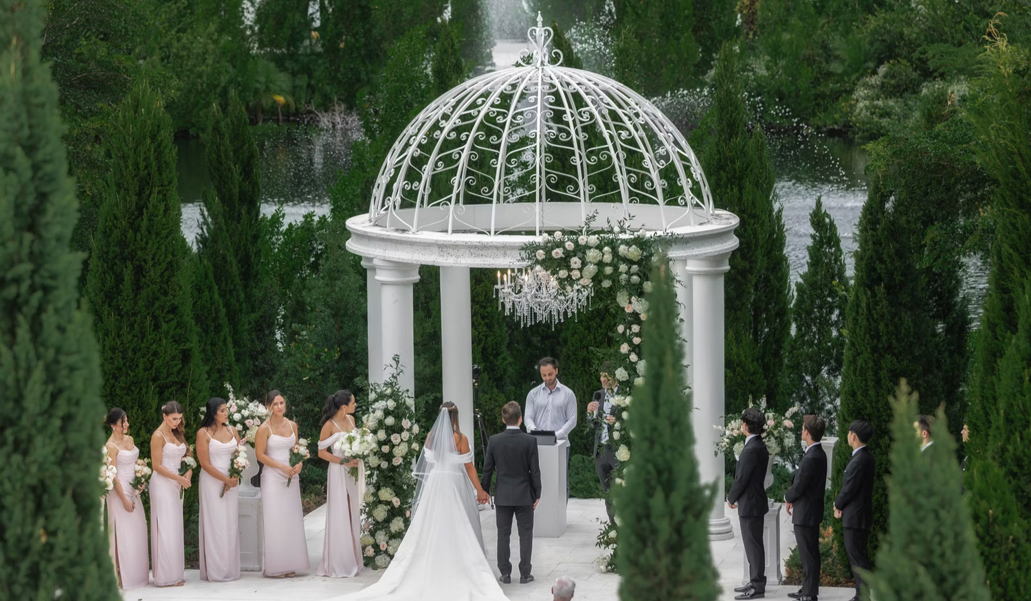 Elegant wedding ceremony takes place under a white domed gazebo surrounded by lush green cypress trees in a garden setting.