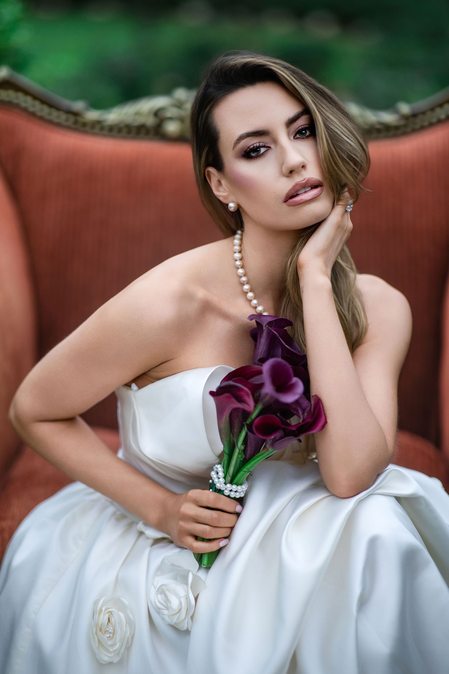 Elegant bride in white strapless gown holding purple calla lily bouquet, seated on vintage red chair wearing pearl necklace.