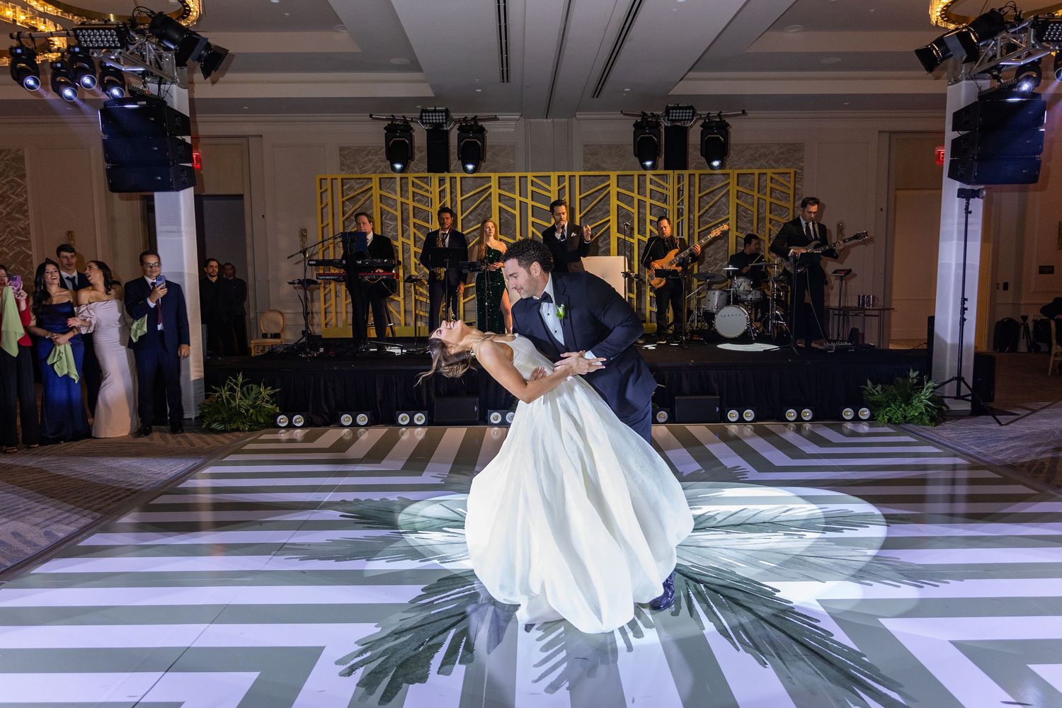Bride and groom share romantic first dance dip on striped dance floor at elegant wedding reception with live band.