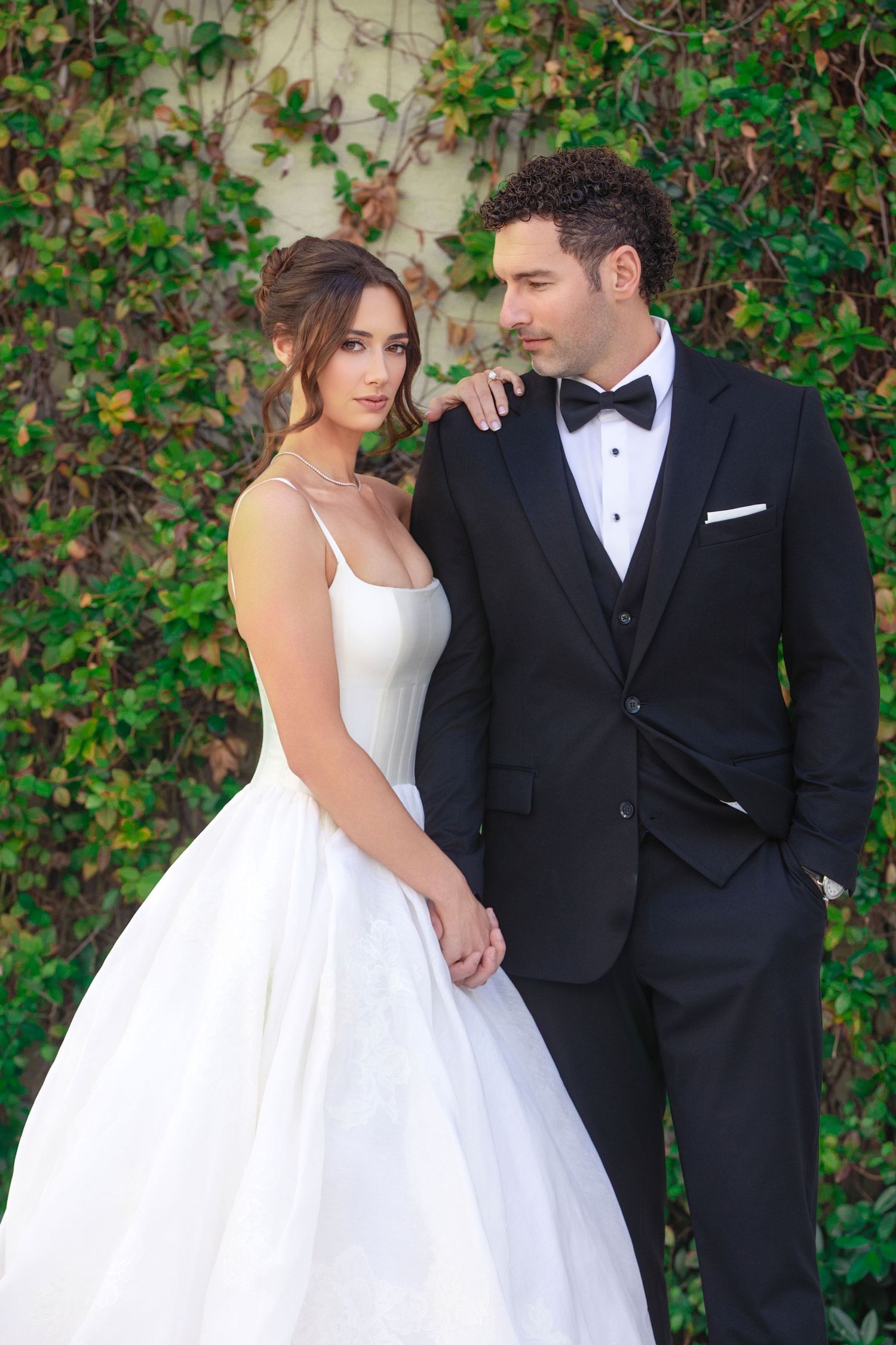 Elegant bride in white ballgown and groom in black tuxedo pose together against ivy-covered wall outdoors.