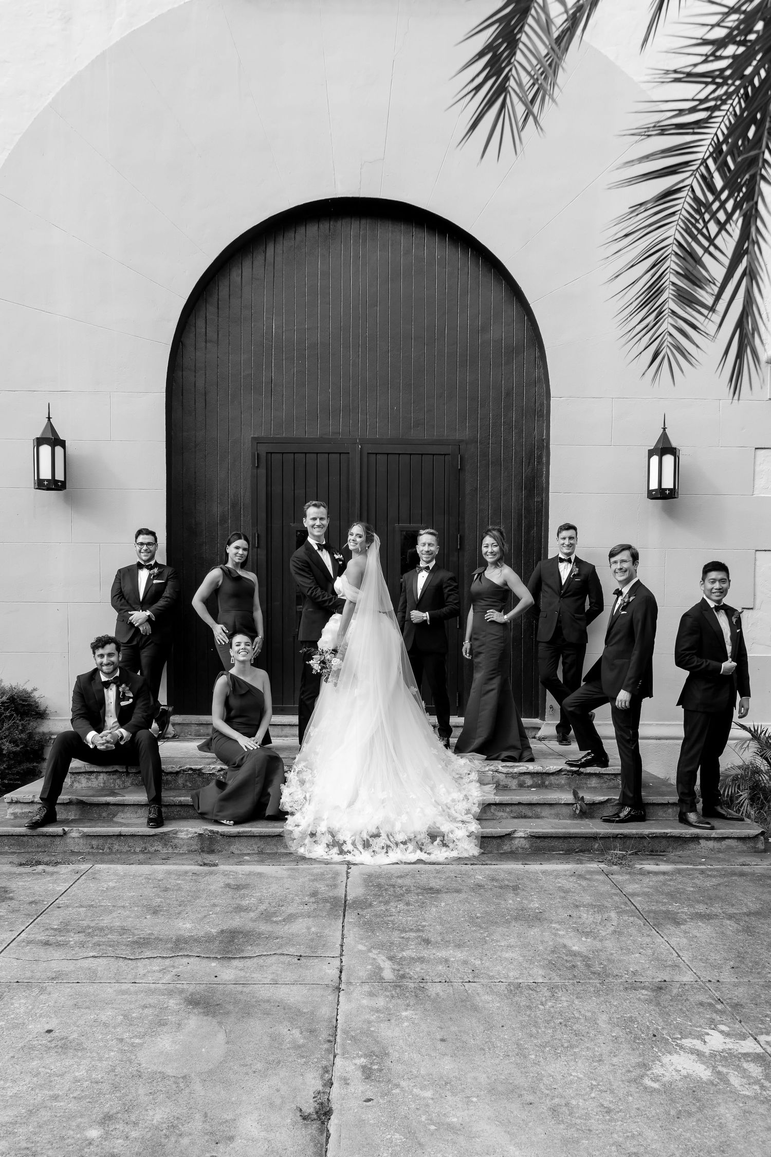 Black and white wedding party photo posed on steps before arched wooden doors with palm fronds and lanterns.