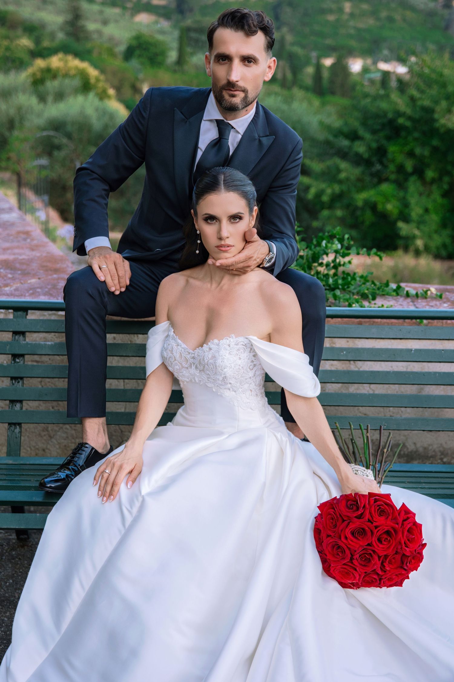 A couple in wedding attire sits on a wooden bench with a vibrant red rose bouquet in an outdoor garden setting.