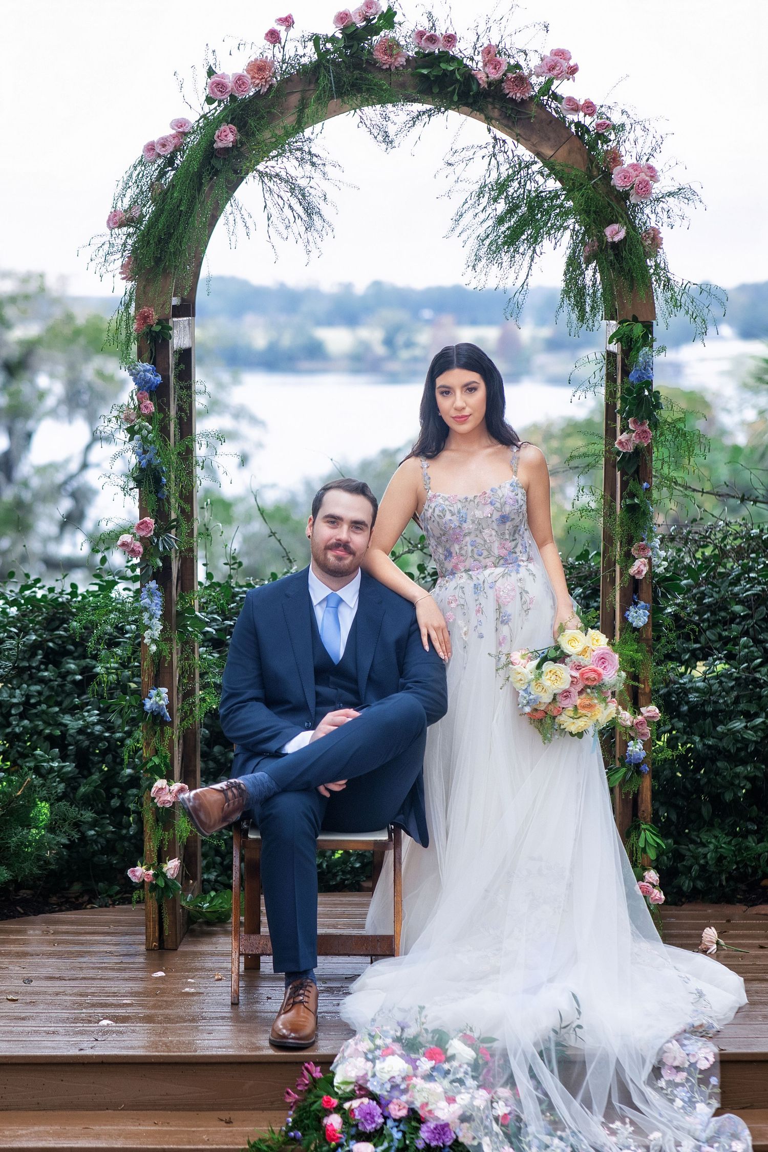 A wedding couple poses under a floral arch decorated with pink roses and greenery on a wooden deck.