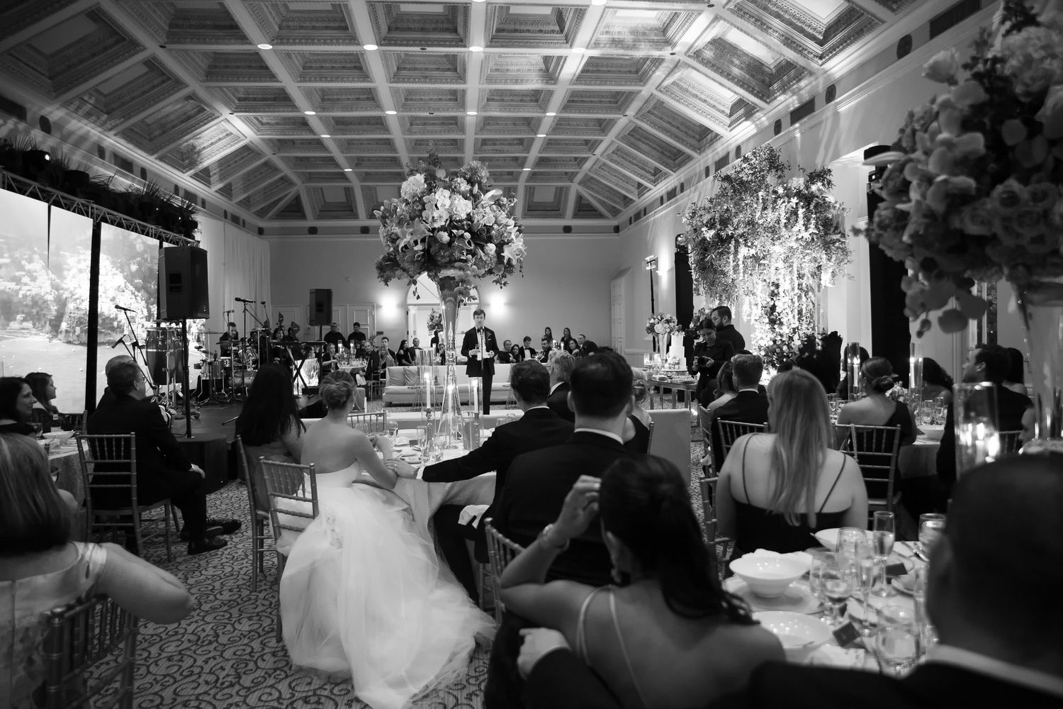 Black and white wedding reception photo showing bride and groom seated among guests in an elegant floral-decorated venue.