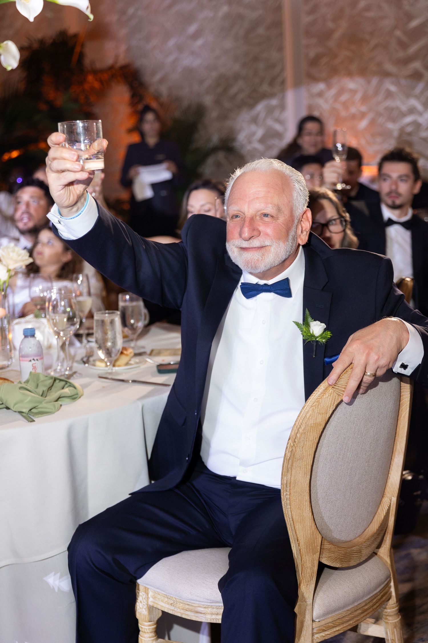 Elderly man in navy tuxedo raises glass for toast at elegant wedding reception, smiling warmly at guests.