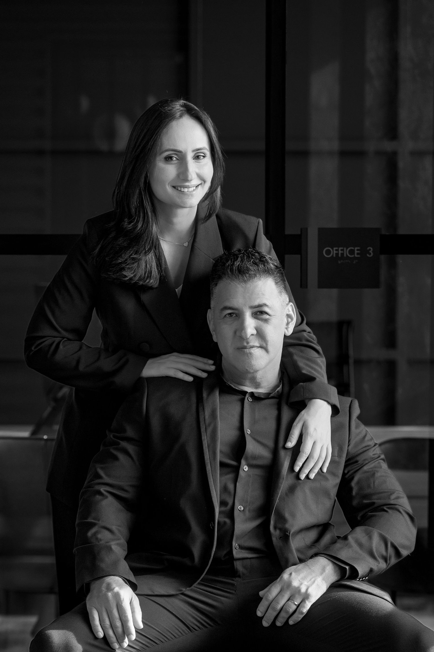 Professional black and white portrait of two people in business attire posing together in an office setting.