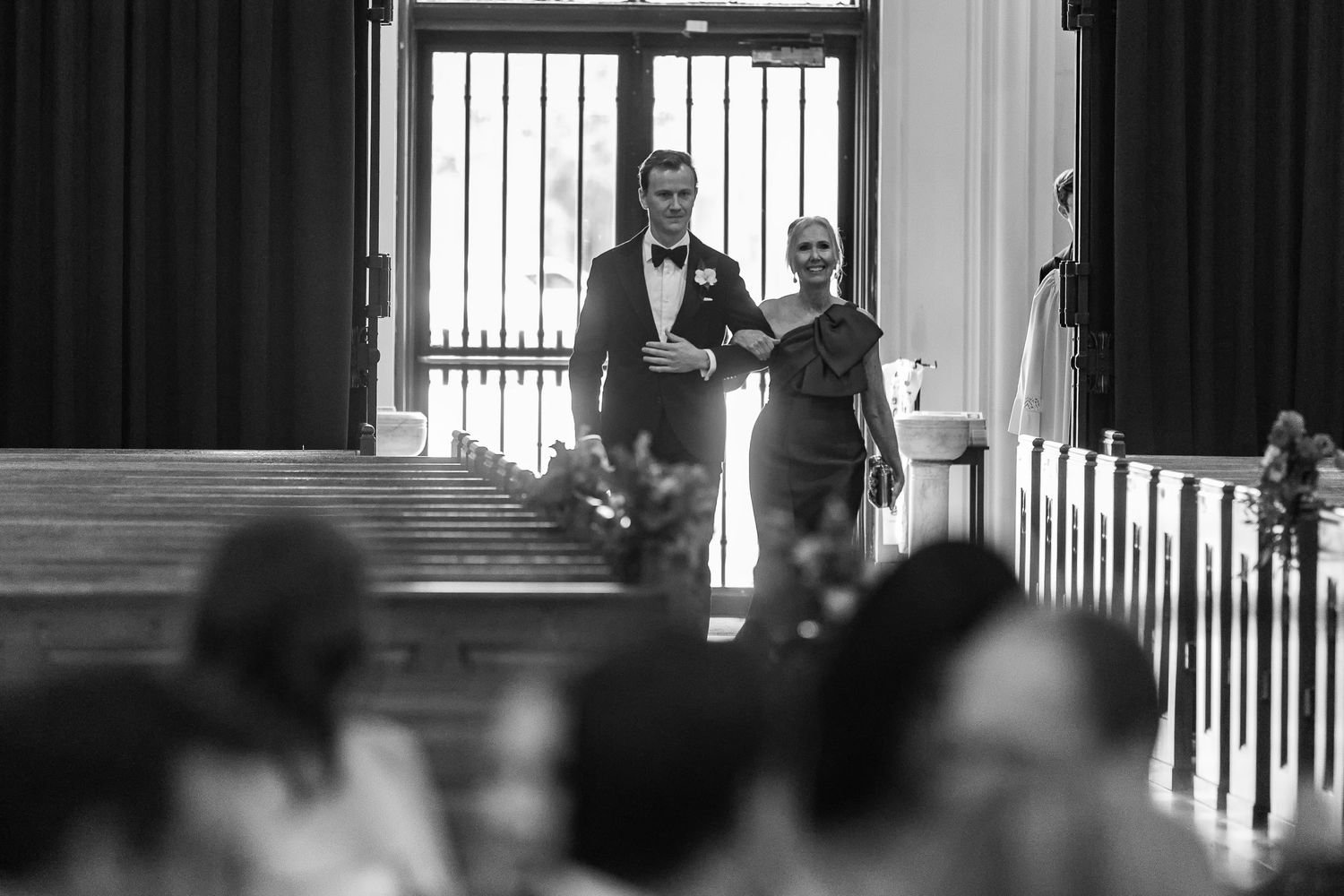 Black and white photo of a groom and bridesmaid walking down church aisle at an elegant wedding ceremony.