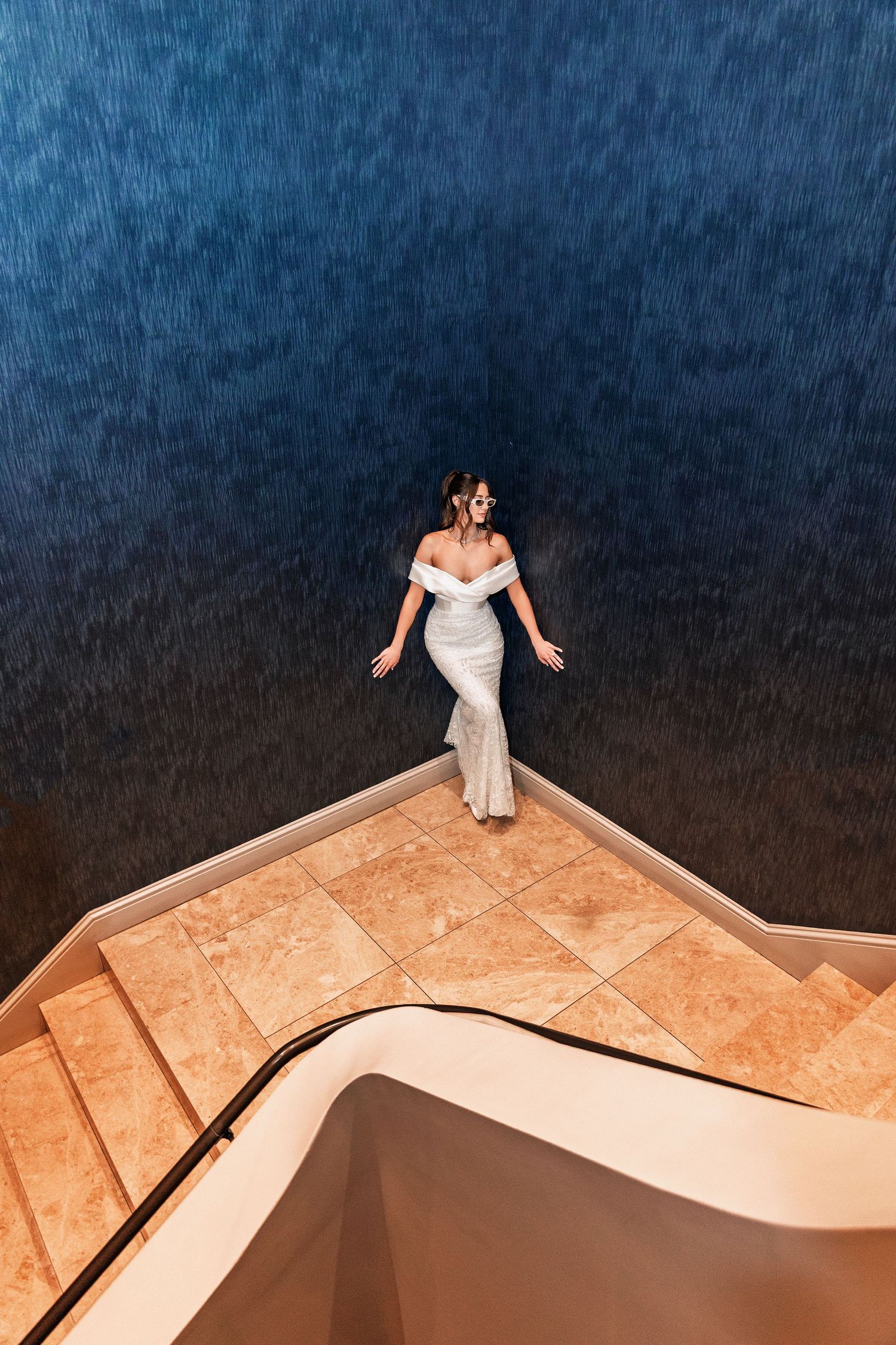 Bride in white off-shoulder gown standing on marble staircase with deep blue textured walls, aerial view.