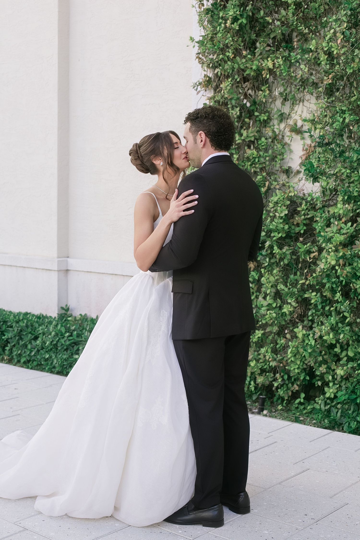 Bride and groom sharing a romantic kiss in front of a lush green ivy wall on their wedding day.