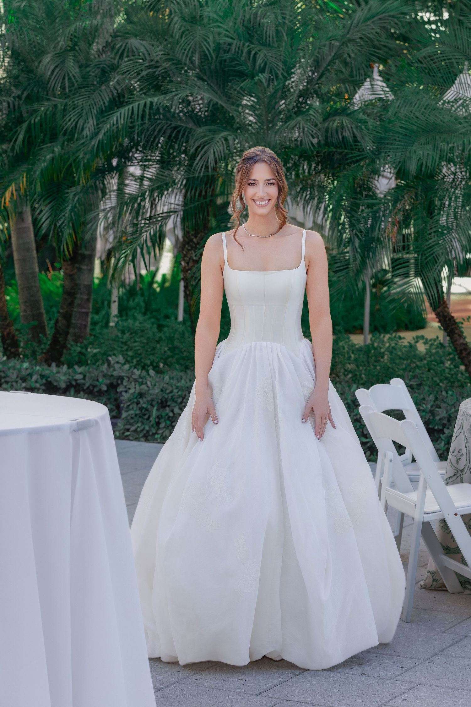 Smiling bride in a white ball gown with spaghetti straps poses outdoors near palm trees and white chairs.