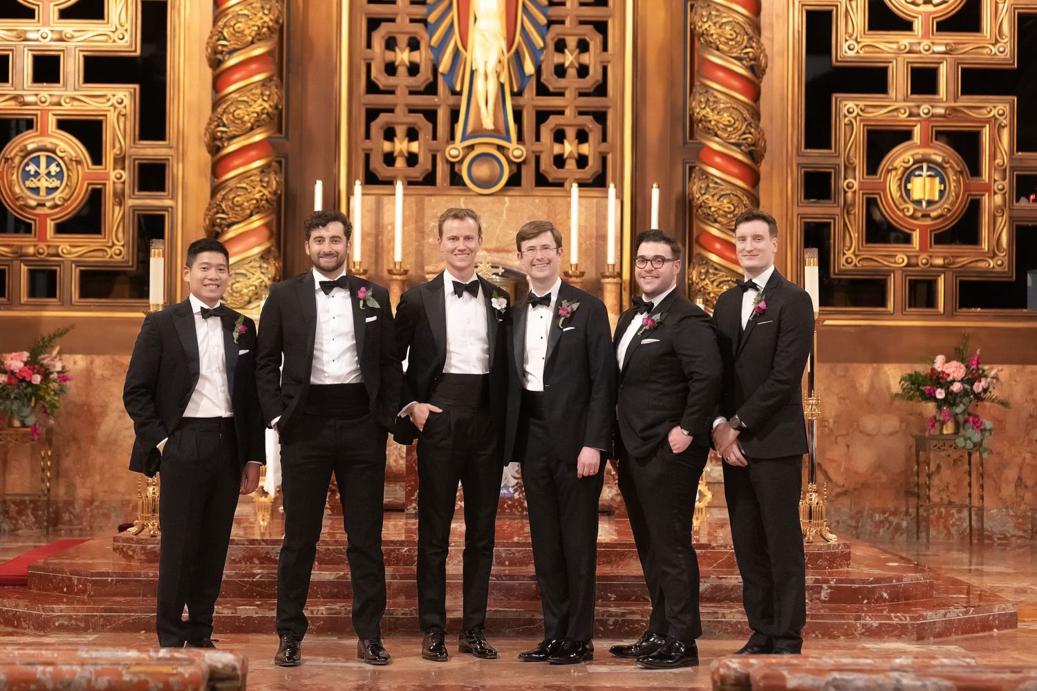 Six groomsmen in black tuxedos posing together at an elegant wedding venue with ornate gold decor.