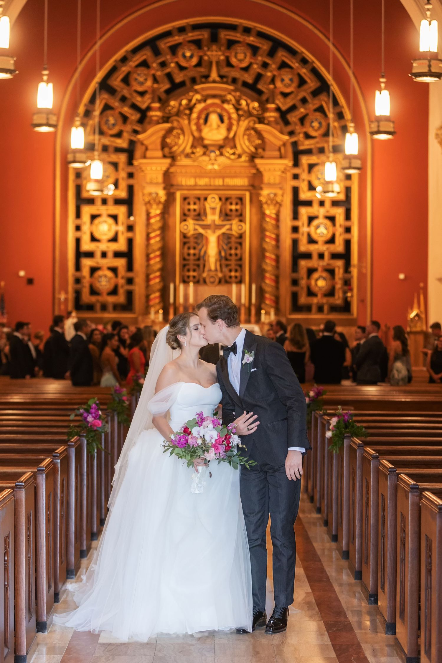 Bride and groom share first kiss in ornate church aisle, bride holding vibrant pink bouquet in elegant white gown.