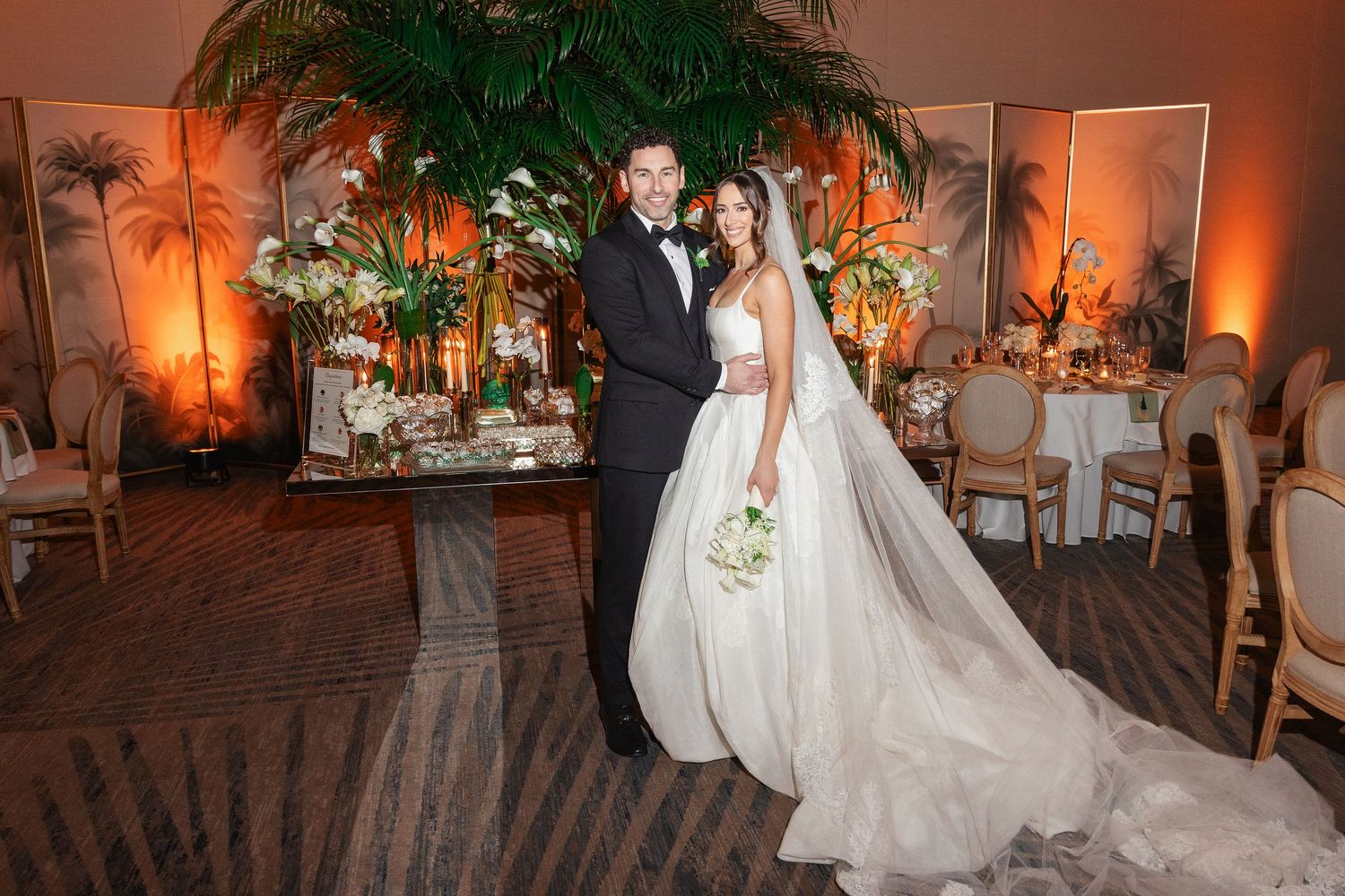 Elegant bride and groom posing in a warmly lit ballroom with lush tropical floral arrangements at their wedding reception.