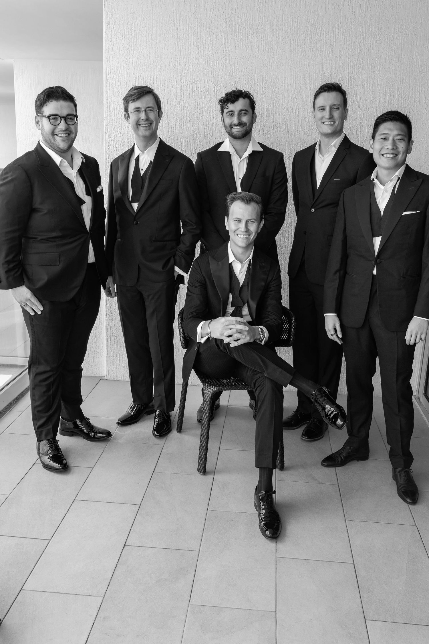 Six men in formal black suits pose together, five standing and one seated on a chair, in a black and white photo.