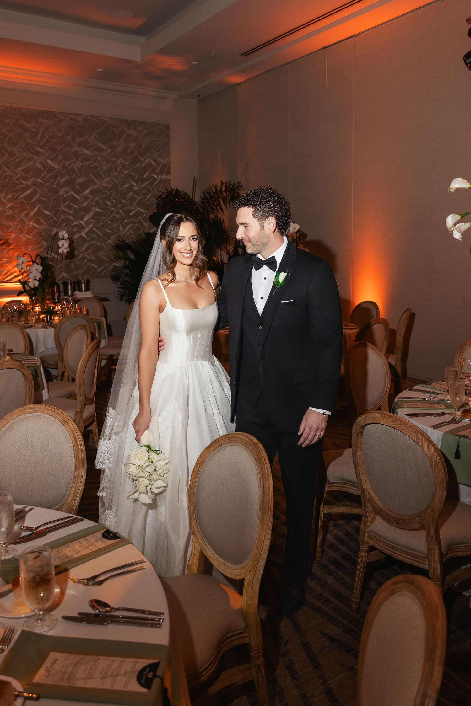 Bride and groom walking through elegant wedding reception venue with warm amber uplighting and gold chairs.