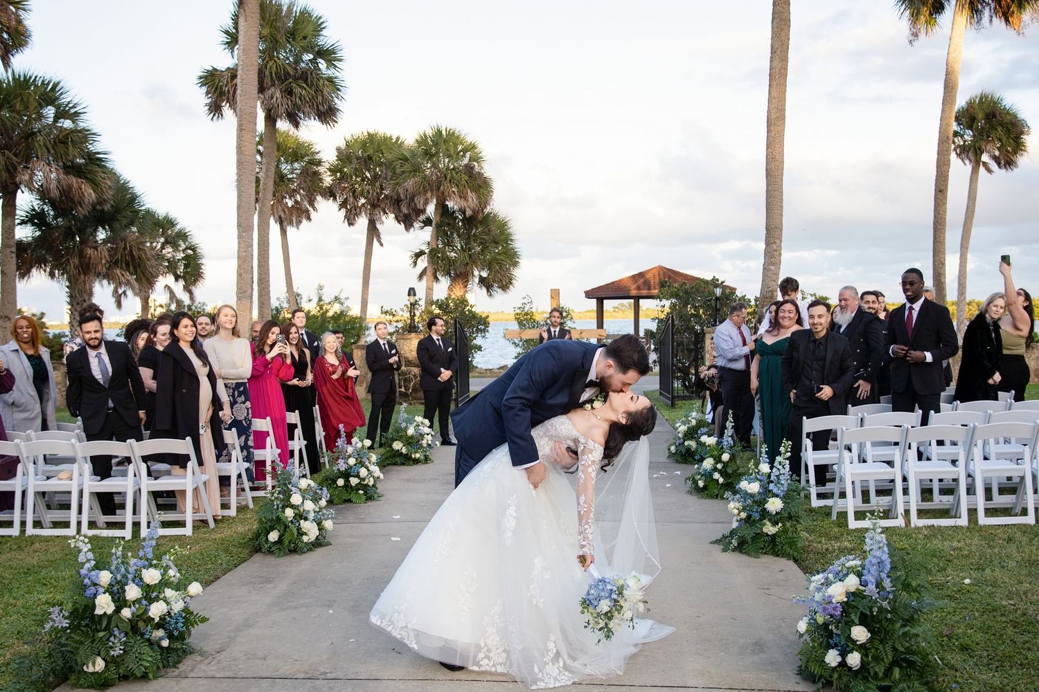 Panoramic view of an outdoor beach wedding ceremony setup with white chairs and palm trees along the coastline.